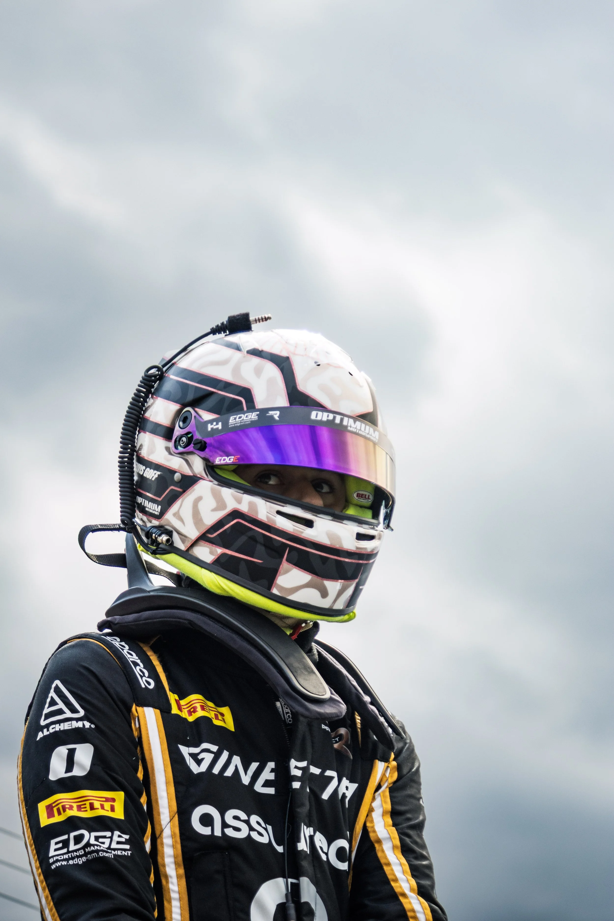 A race car driver wearing a black racing suit with sponsor logos, a full-face helmet with a reflective visor, standing outdoors against cloudy skies.