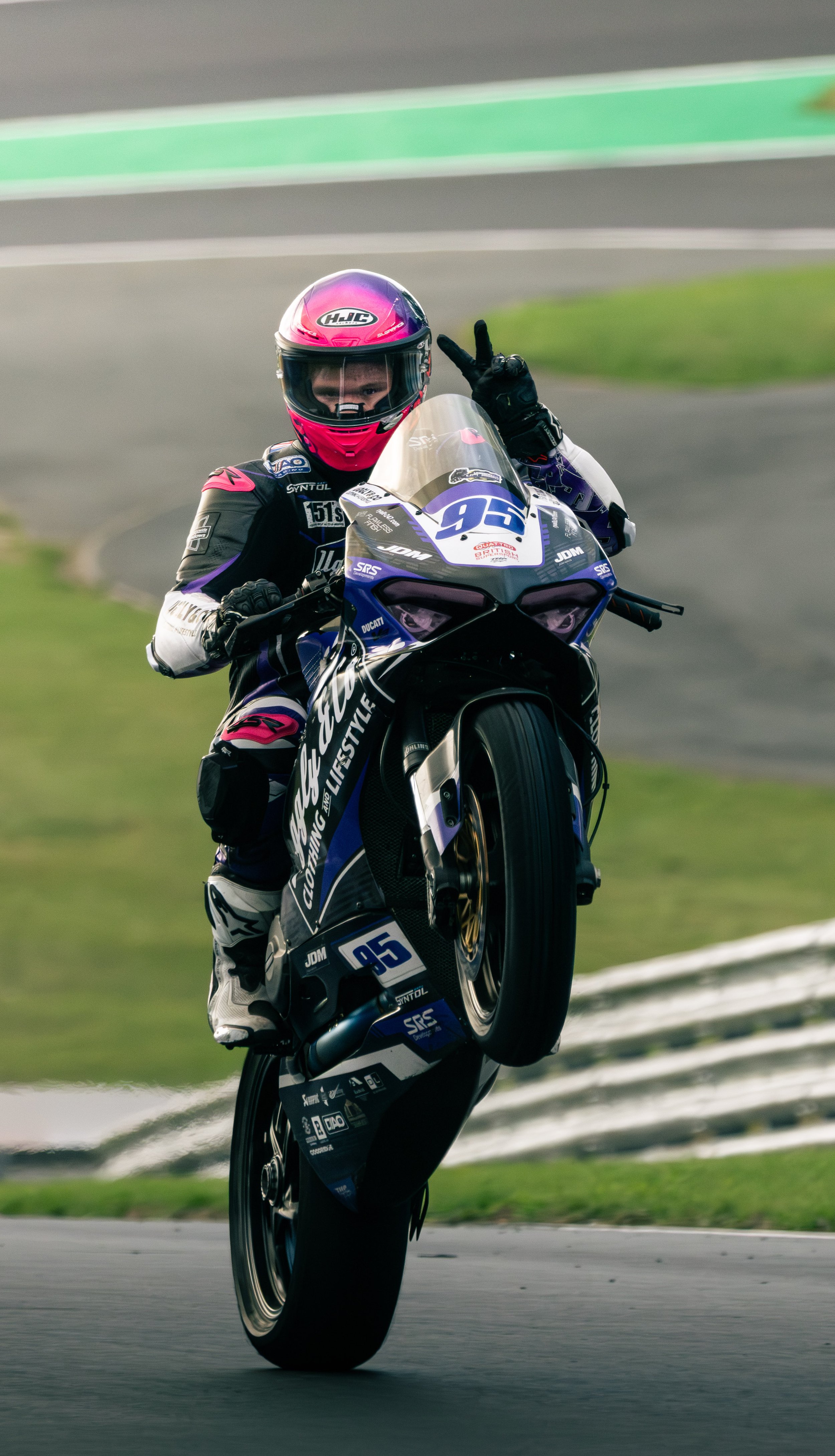 A motorcycle racer wearing a pink helmet performs a wheelie on a racetrack, giving a peace sign with his right hand.