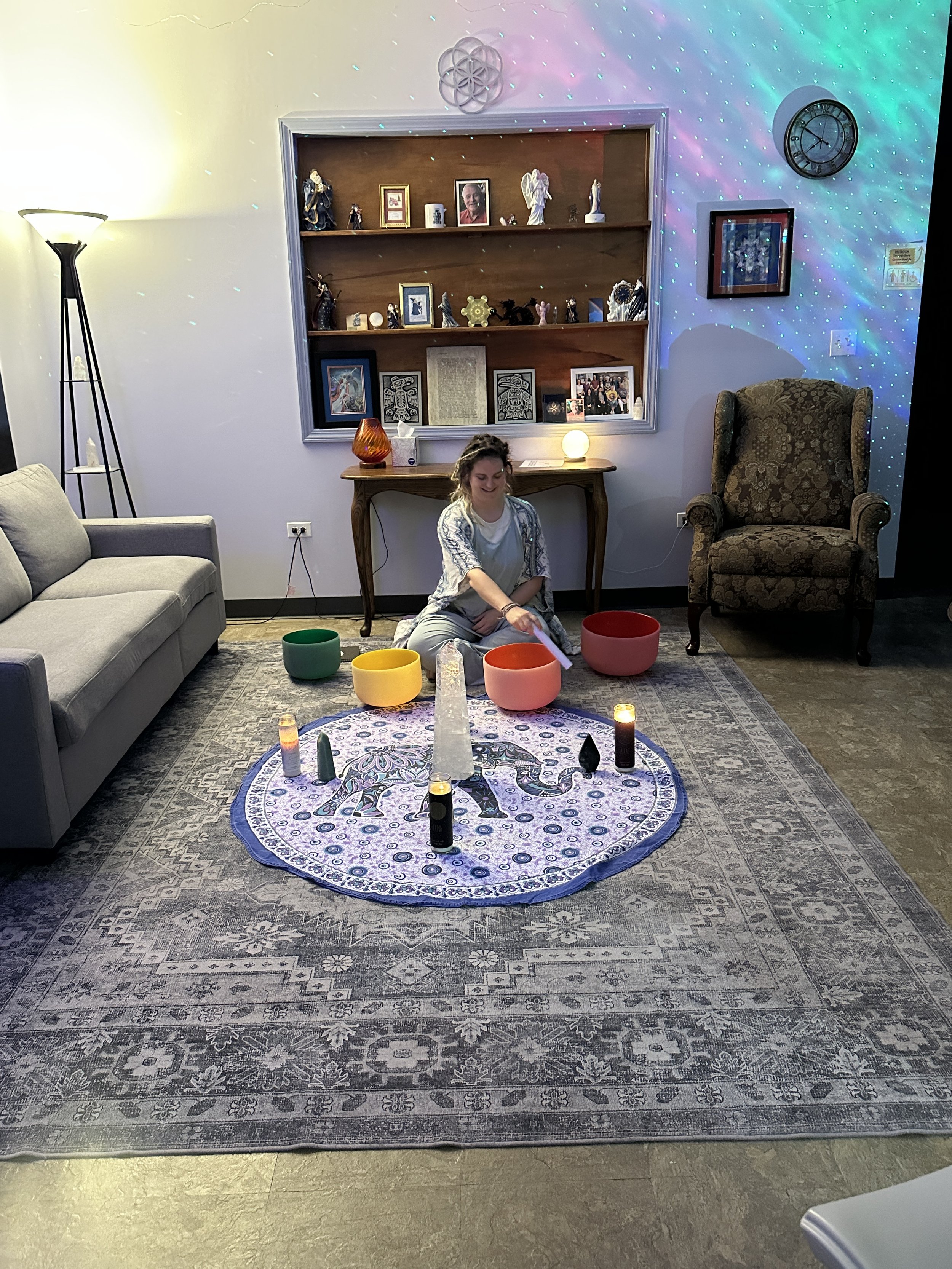 A woman sitting on the floor in front of a decorated altar with candles and bowls, a patterned rug, a couch on the left, a chair on the right, a wooden shelf with statues and photographs on the back wall, a tall lamp, and a clock on the wall.