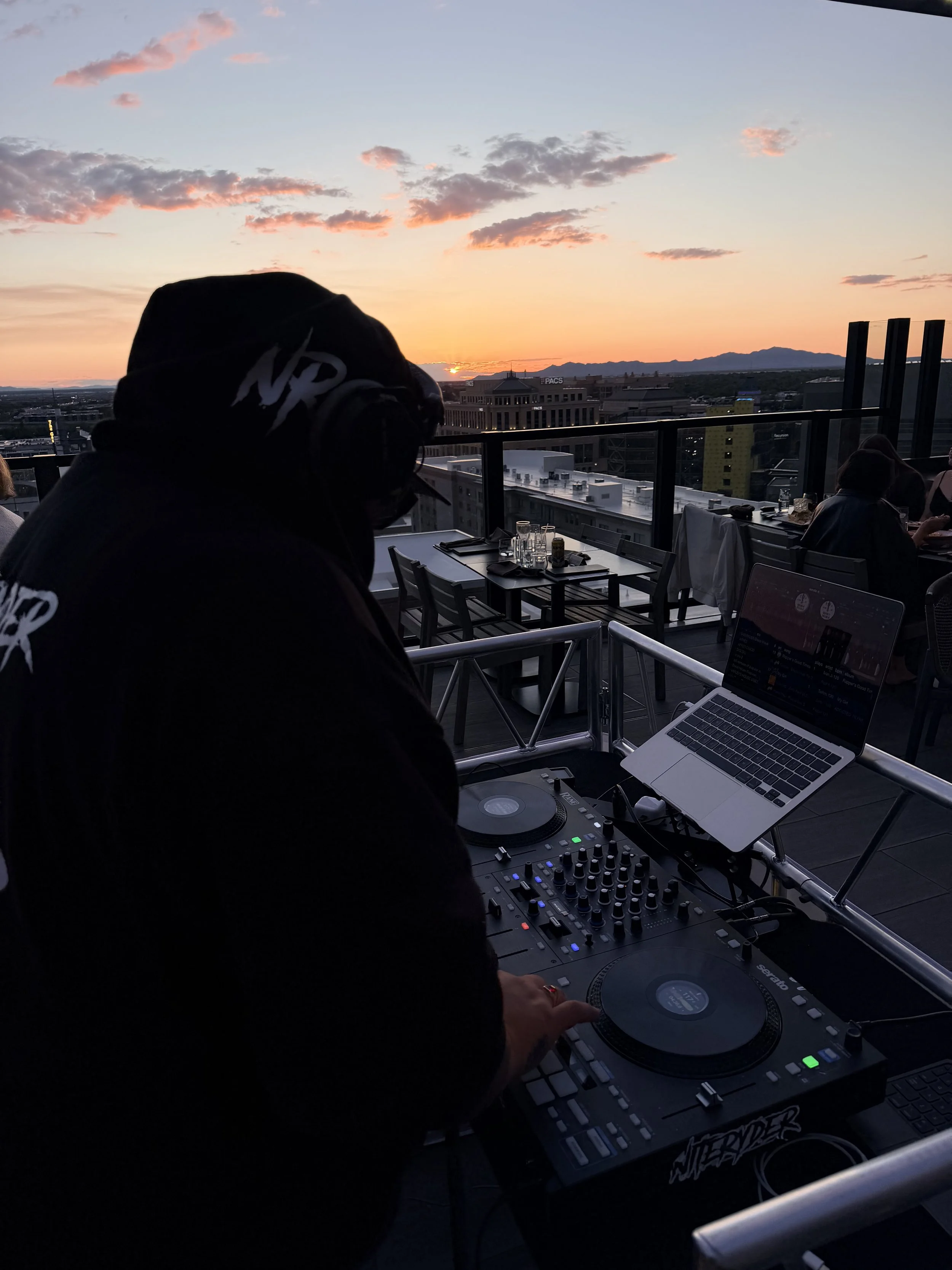 A DJ wearing headphones and a hoodie is operating DJ equipment with a cityscape and a sunset in the background.