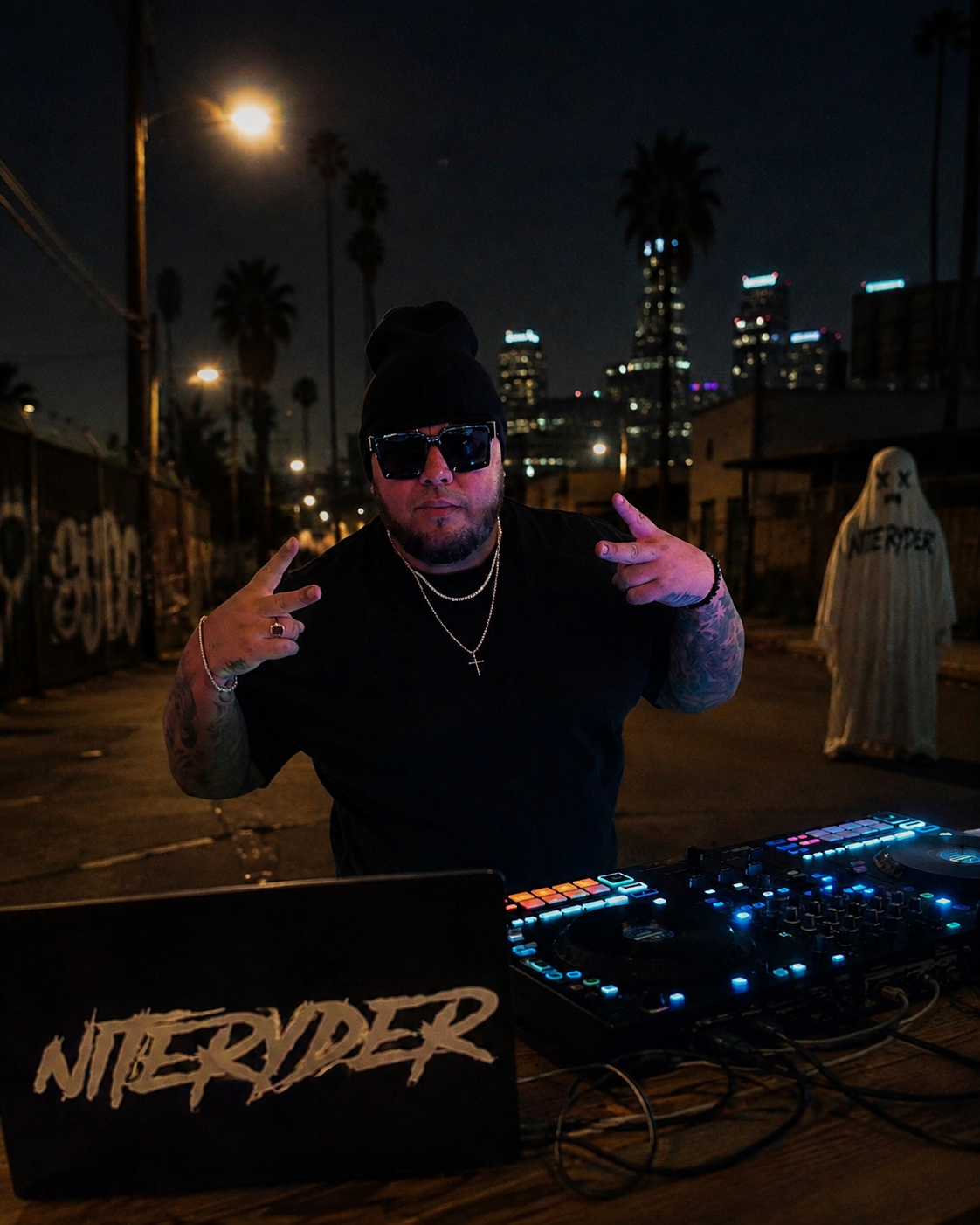 A man DJing at night outdoors with city skyscrapers in the background. He wears black sunglasses, a black beanie, and jewelry, and is posing with both hands making peace signs. There's a DJ mixer and laptop with a Niterider sticker on the table.