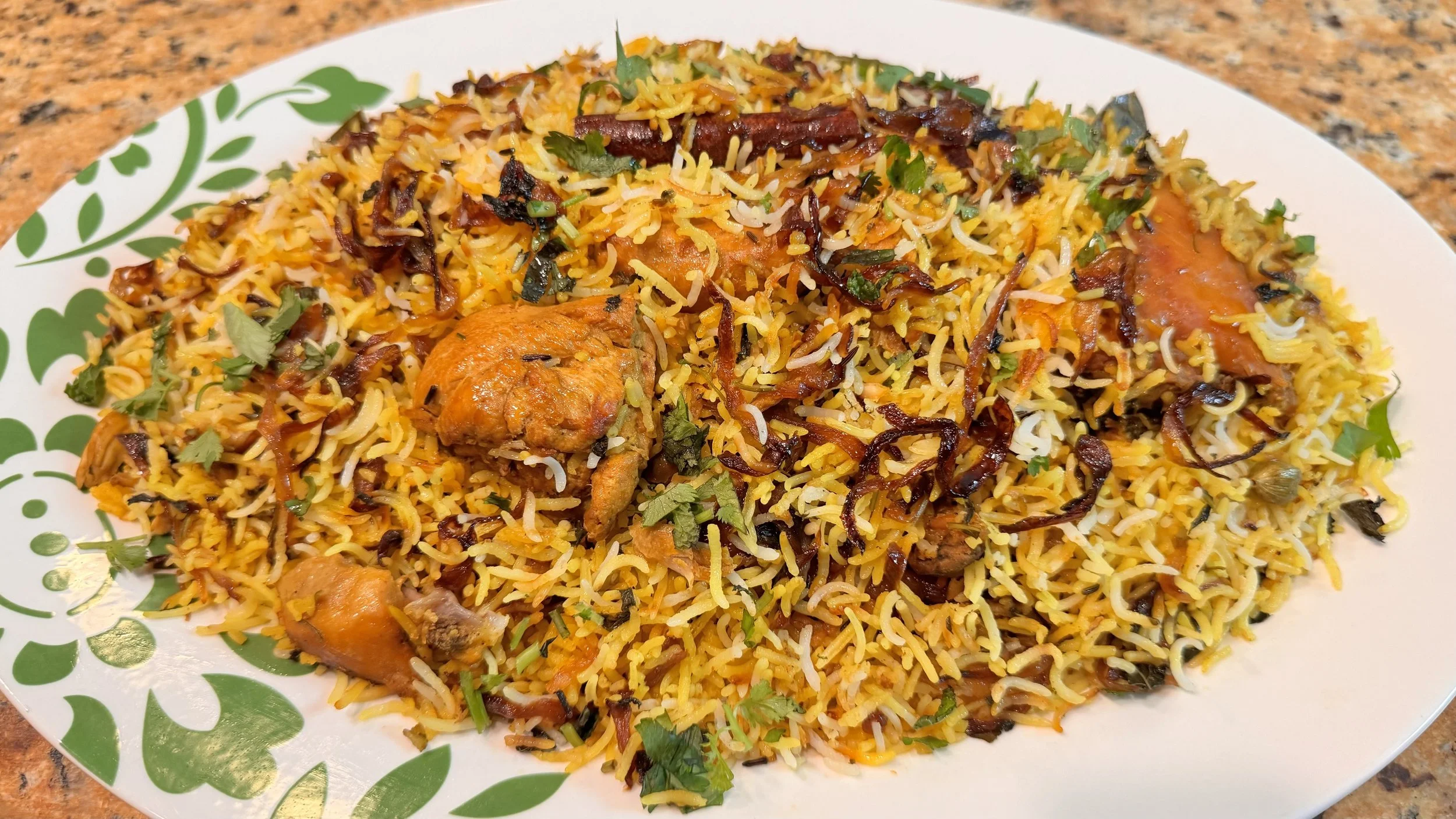 A plate of cooked biryani rice garnished with herbs, with pieces of cooked chicken and garnishes of herbs and fried onions.
