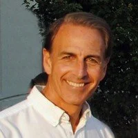 A smiling man with light skin and brown hair pulled back, wearing a white collared shirt, standing outdoors with trees in the background.