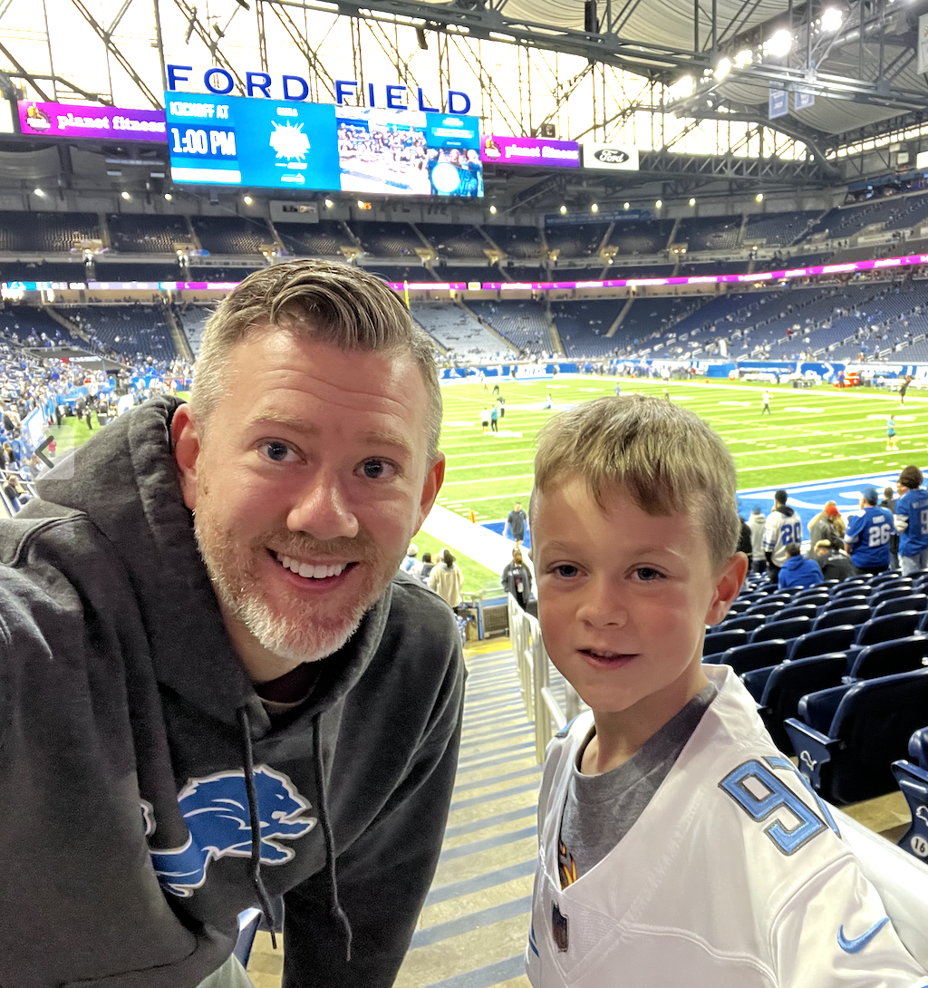 A man and a young boy taking a selfie inside Ford Field stadium during a football game. The stadium is partially filled with spectators, and the field is being prepared for play.
