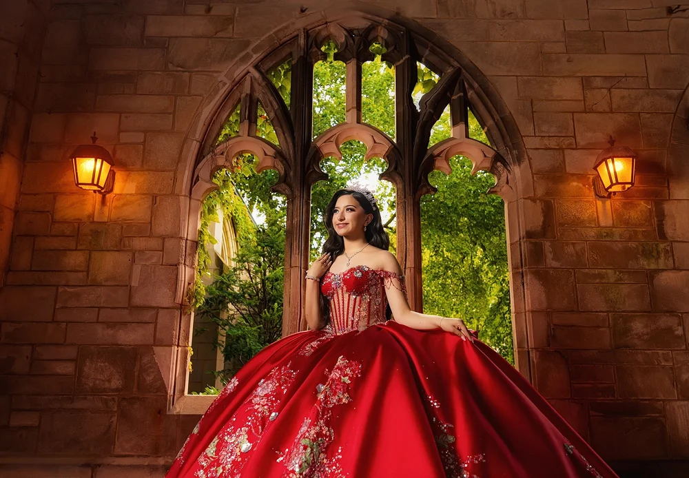 Genesis in a red quinceañera gown beneath a Gothic stone arch with warm lantern light
