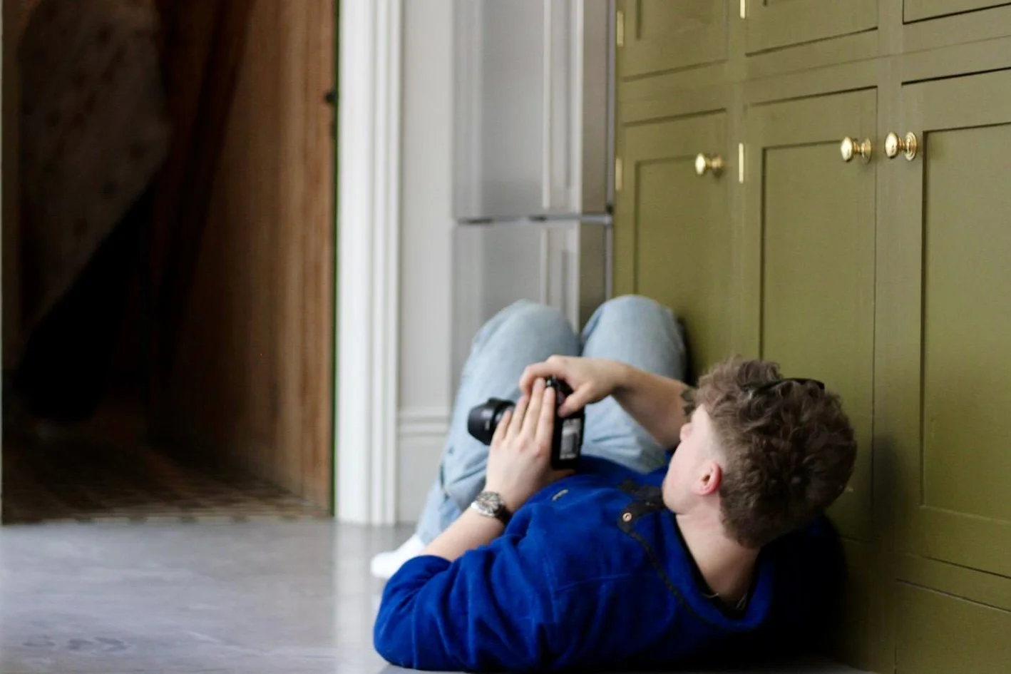 A young man with curly hair wearing a blue hoodie and jeans lies on the wooden floor, leaning against green kitchen cabinets, taking a photo with a camera.