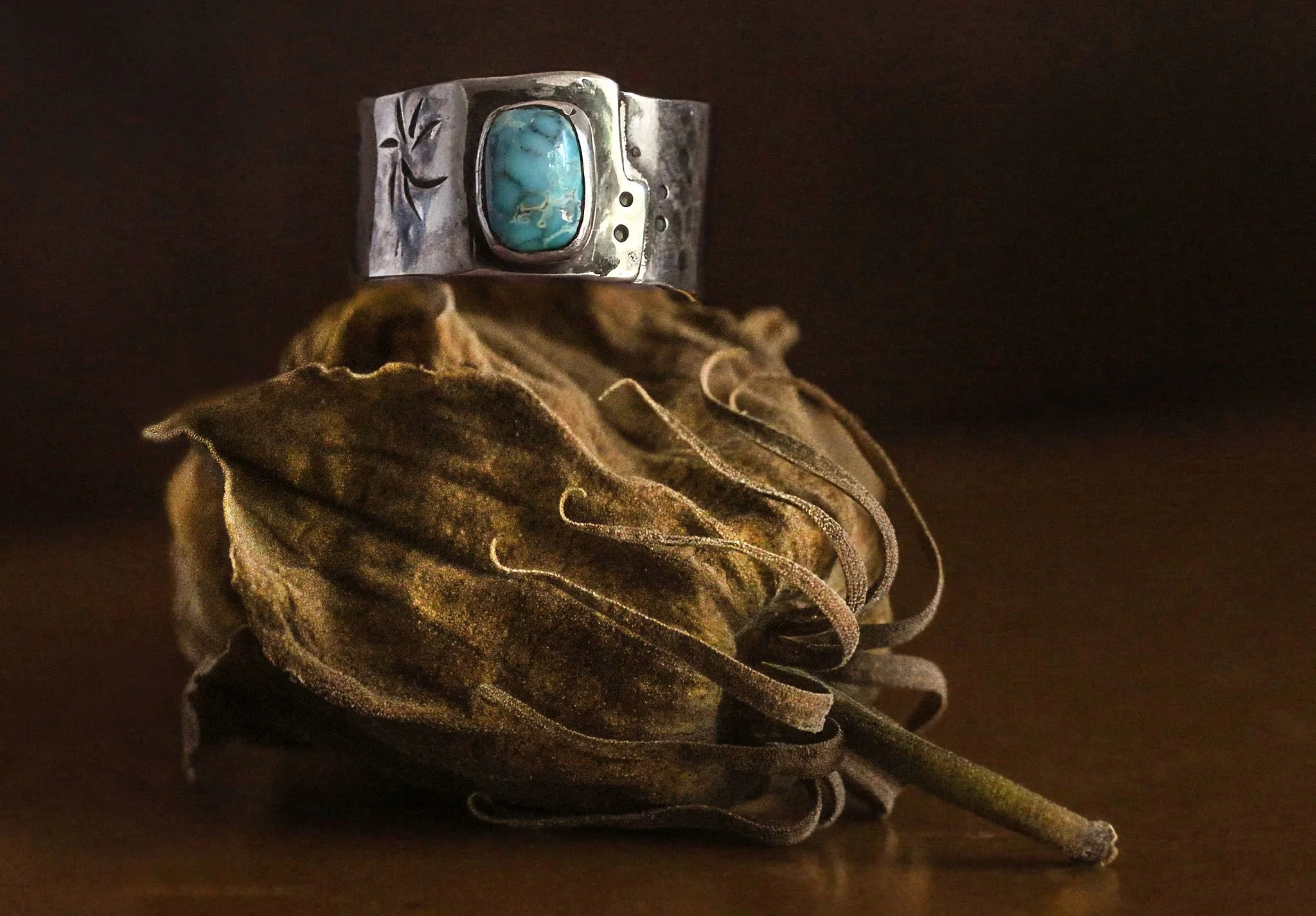 Artistic still life photograph featuring a handcrafted sterling silver cuff bracelet with a turquoise stone, styled on a dried botanical pod. This moody, textured composition highlights natural materials, earthy tones, and artisan jewelry design. Ide