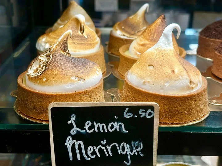 Lemon meringue tarts displayed in a bakery case, with a chalkboard sign showing the price of 6.50.
