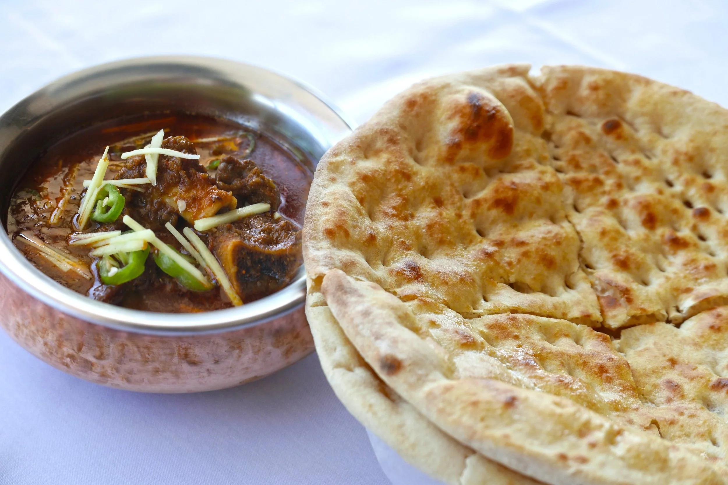 A bowl of Indian curry garnished with shredded cheese and chopped green chili, served next to a piece of naan bread.