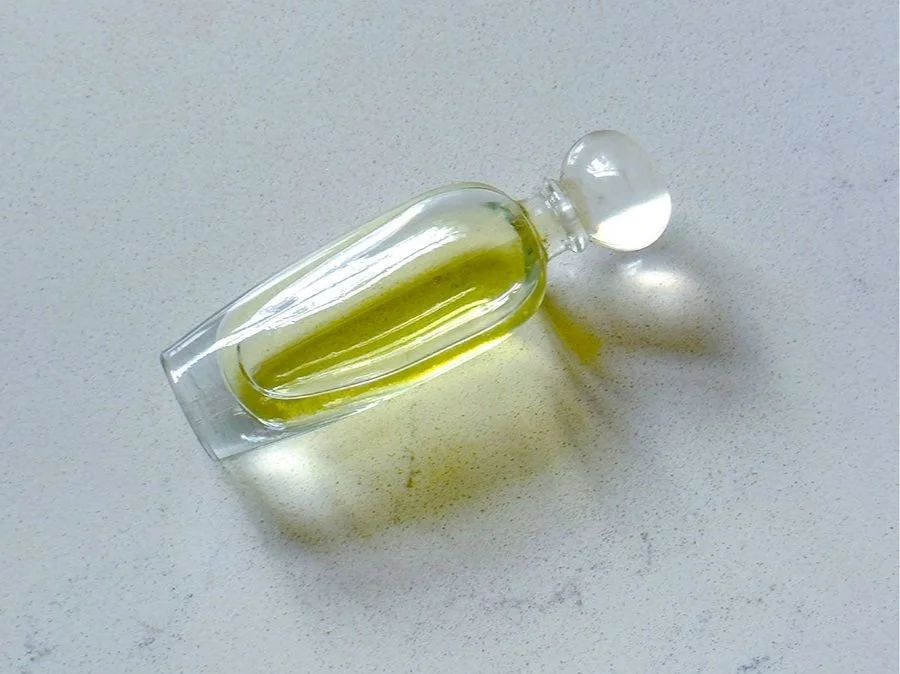 A small glass vial with a rounded glass stopper, filled with a yellow-green liquid, laying on a white surface with some liquid spilled nearby.