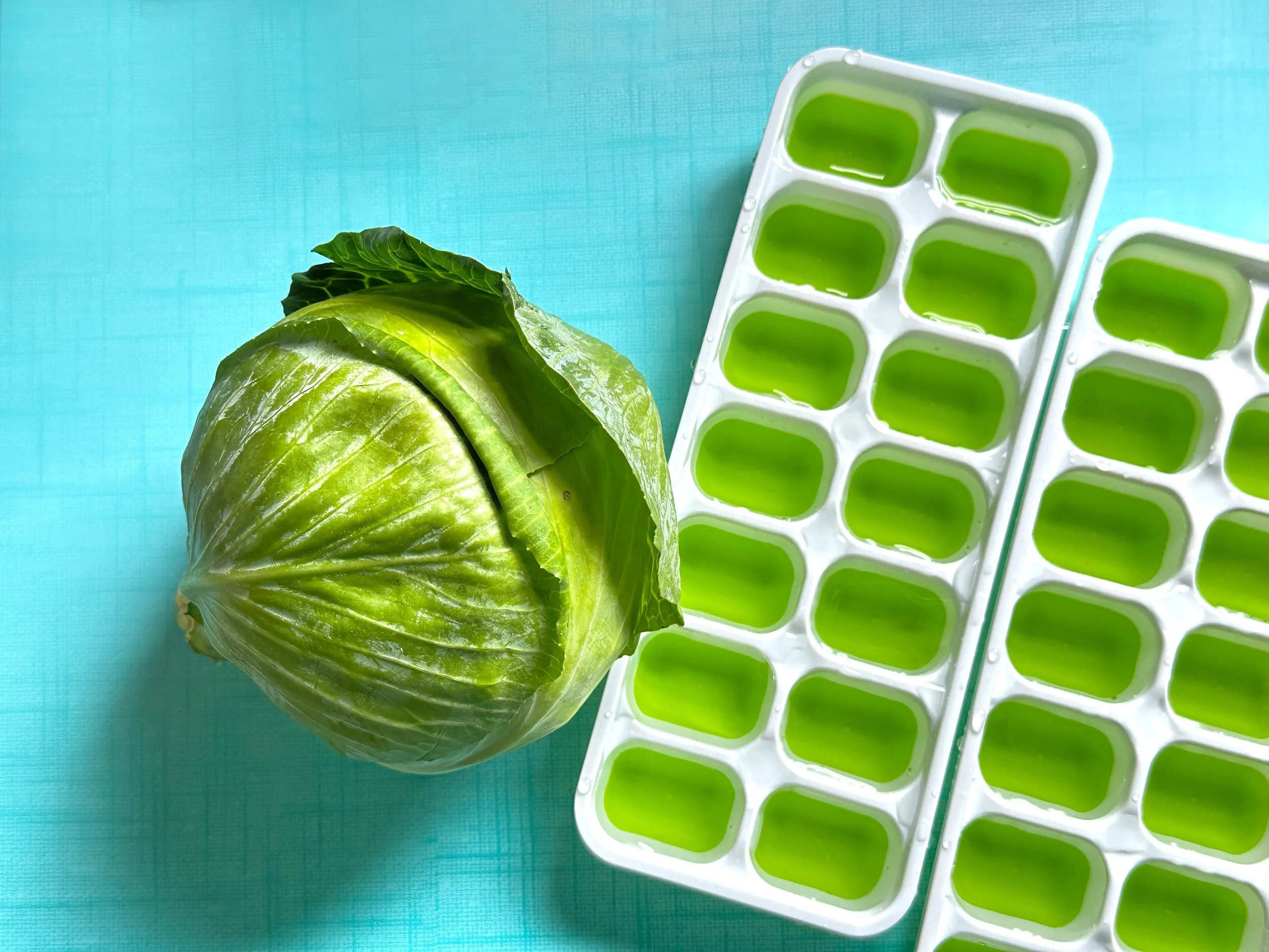 A head of fresh green lettuce next to two white ice cube trays filled with green liquid on a light blue surface.