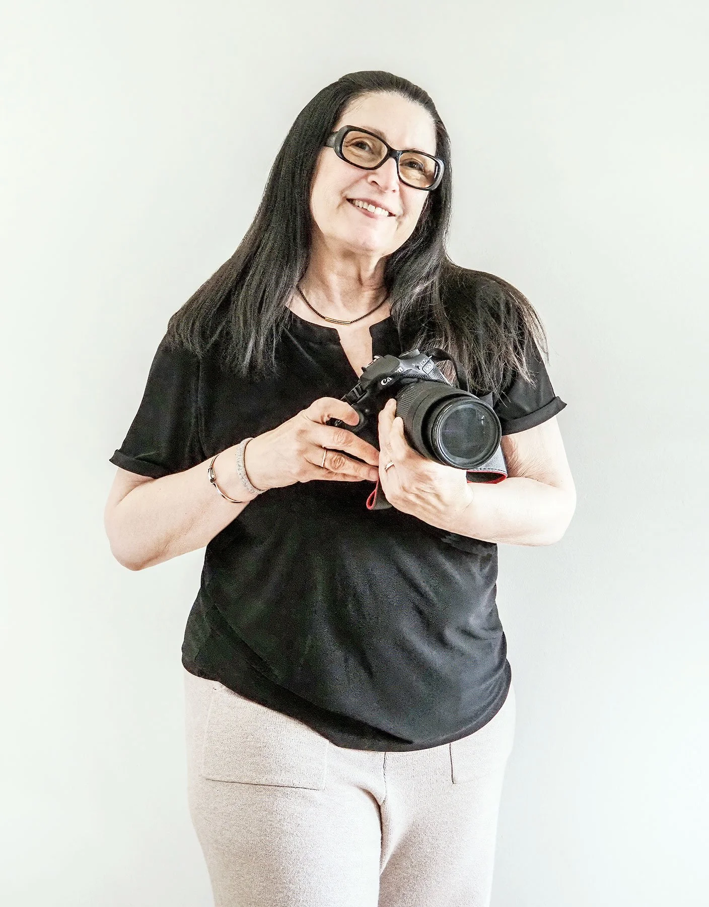 A woman with long dark hair, glasses, and a black shirt holding a DSLR camera, smiling at the camera. She is standing against a plain, light-colored background.