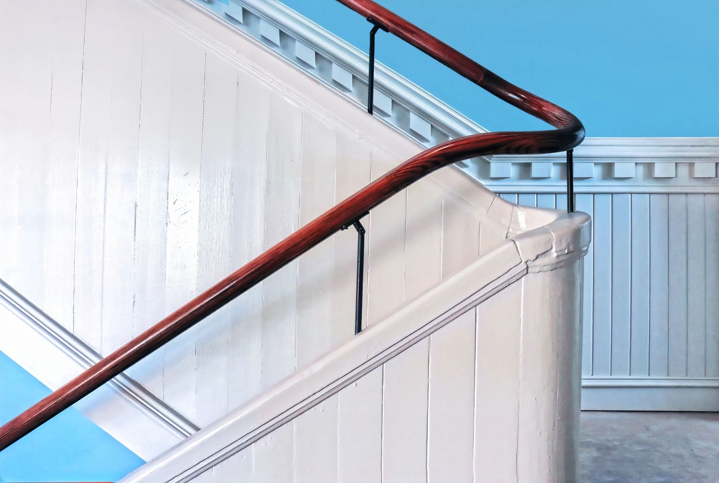 Interior photograph of a staircase with white wainscoting, a dark wooden handrail, and a blue wall.
