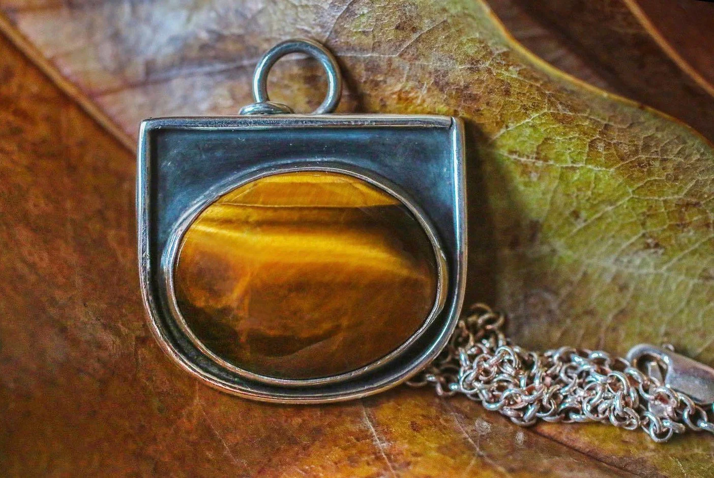 A pendant with a tiger's eye stone in a silver setting on a chain, resting on a brown leaf.