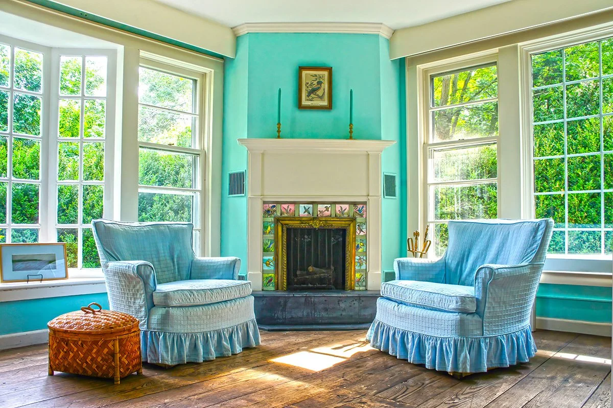  Interior photography of a living room shows large windows, light blue walls, a white fireplace, and two matching light blue armchairs.