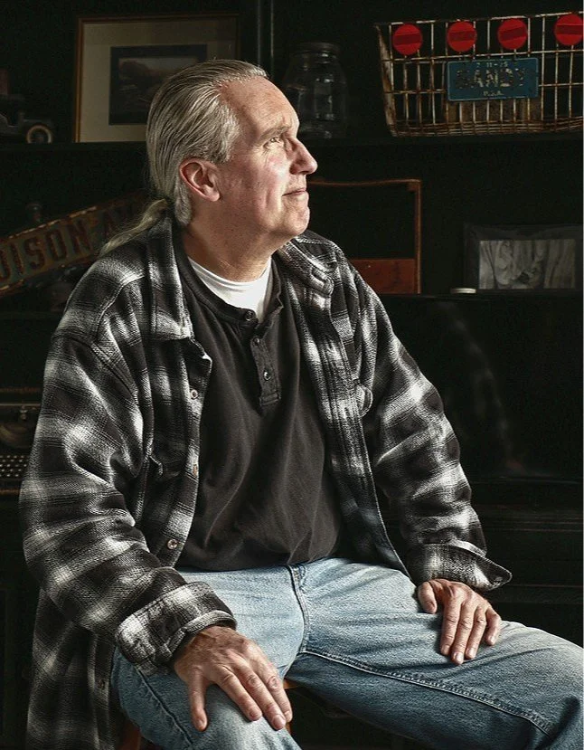 Older man with gray hair sitting on a chair, wearing a black and white flannel shirt over a black T-shirt and light blue jeans, looking off to the side in a dimly lit room.