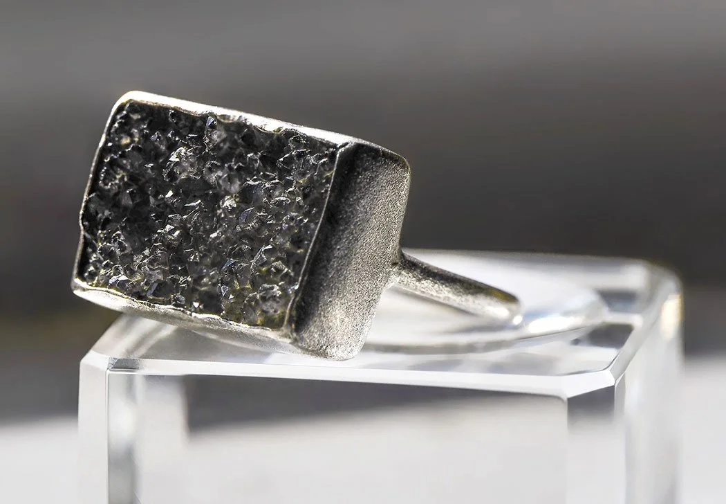 A close-up of a silver ring with a rectangular black druzy gemstone, displayed on a clear stand.