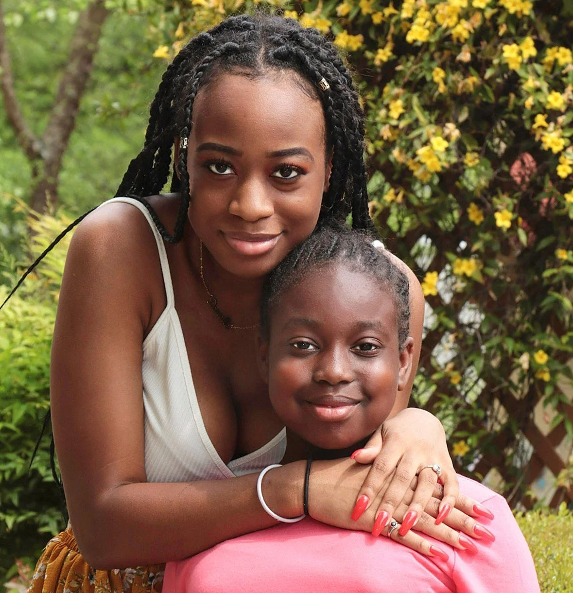 A young woman with braided hair and a young girl with braided hair smiling outdoors, surrounded by greenery and yellow flowers.