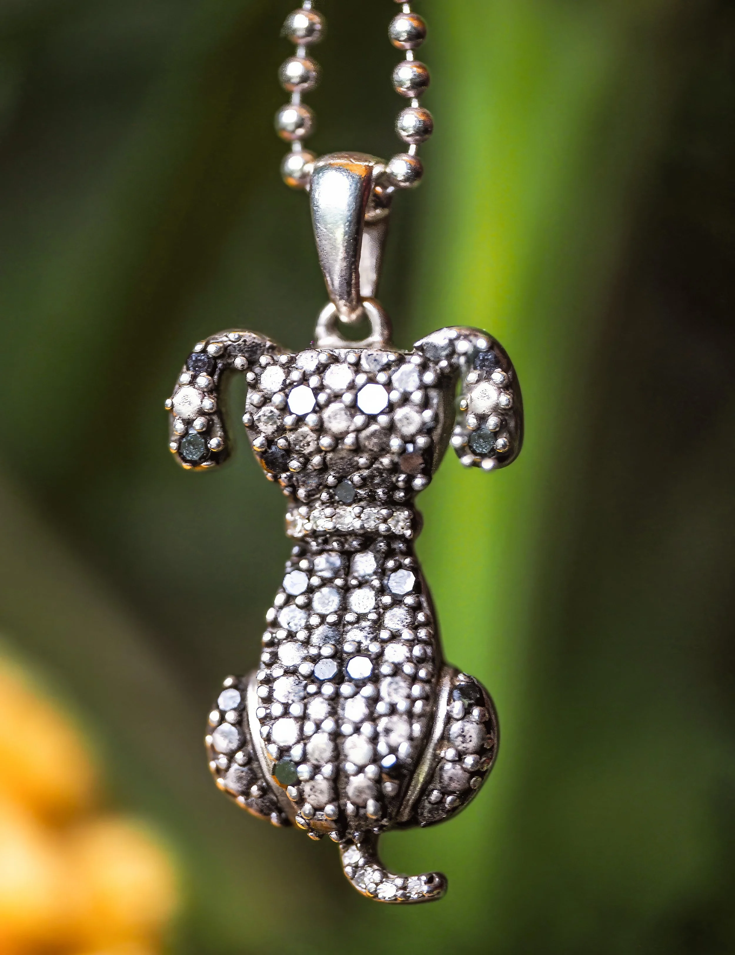 A close-up of a silver dog-shaped pendant necklace decorated with numerous small rhinestones, hanging from a beaded chain, is a product photograph by Margaret Waage Photography.