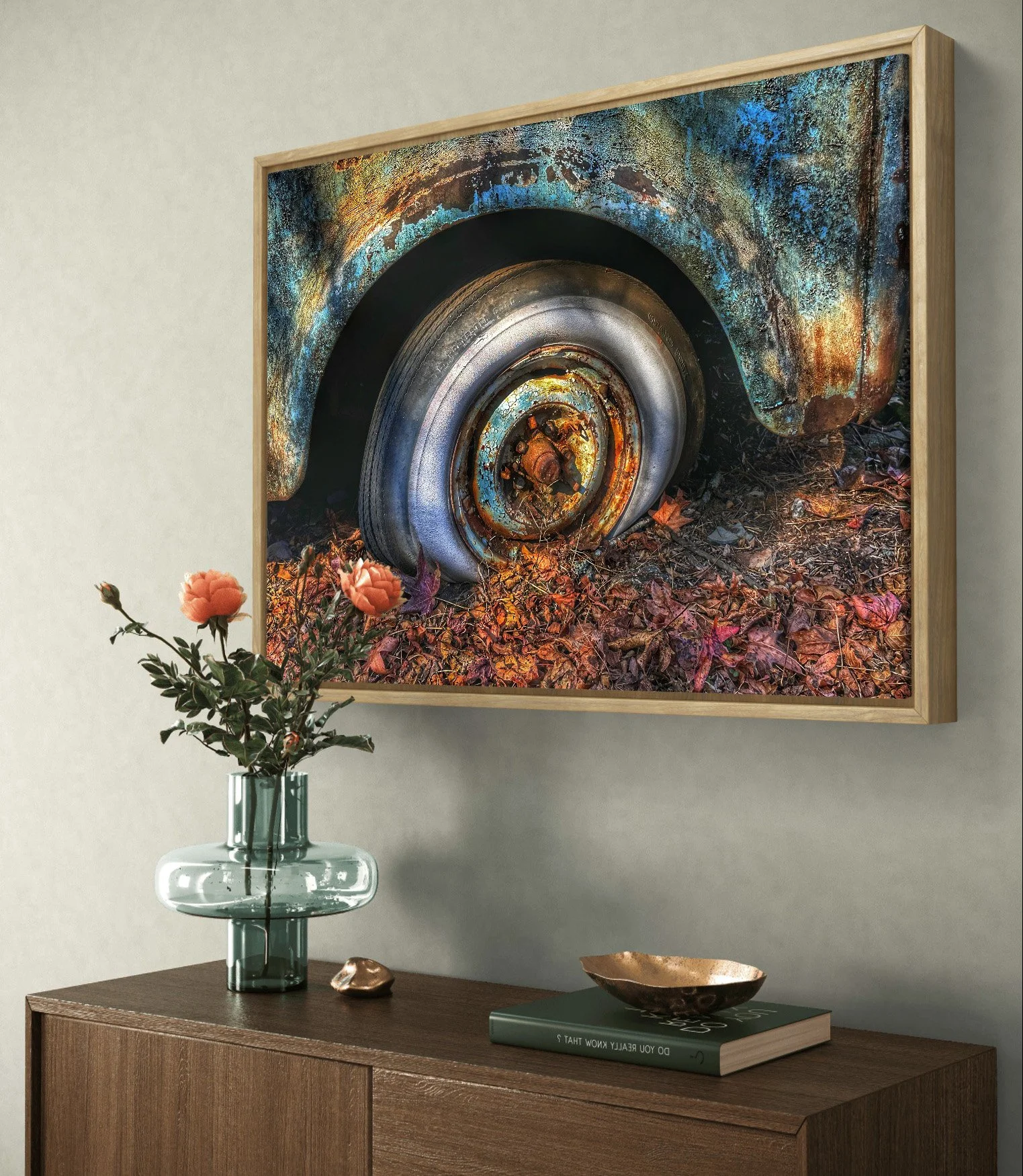 A framed photograph of a rusty, abandoned car wheel on leaf-covered ground is hung on a wall. A wooden sideboard below has a green vase with peach-colored flowers, a decorative rock, a green book, and a small metallic bowl.