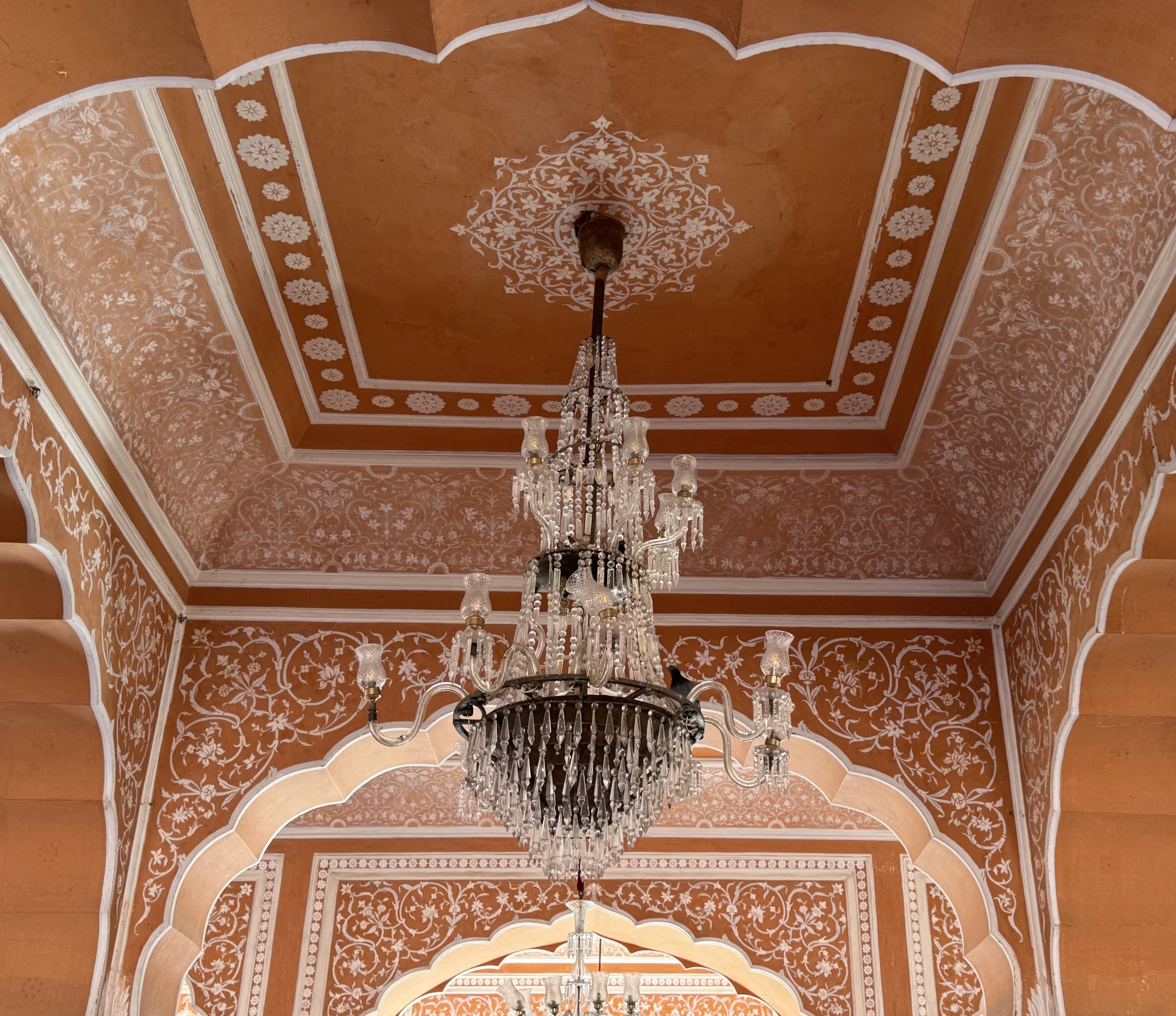 View of an ornate ceiling with decorative white floral and vine patterns on a warm orange background and a large crystal chandelier hanging from the center.