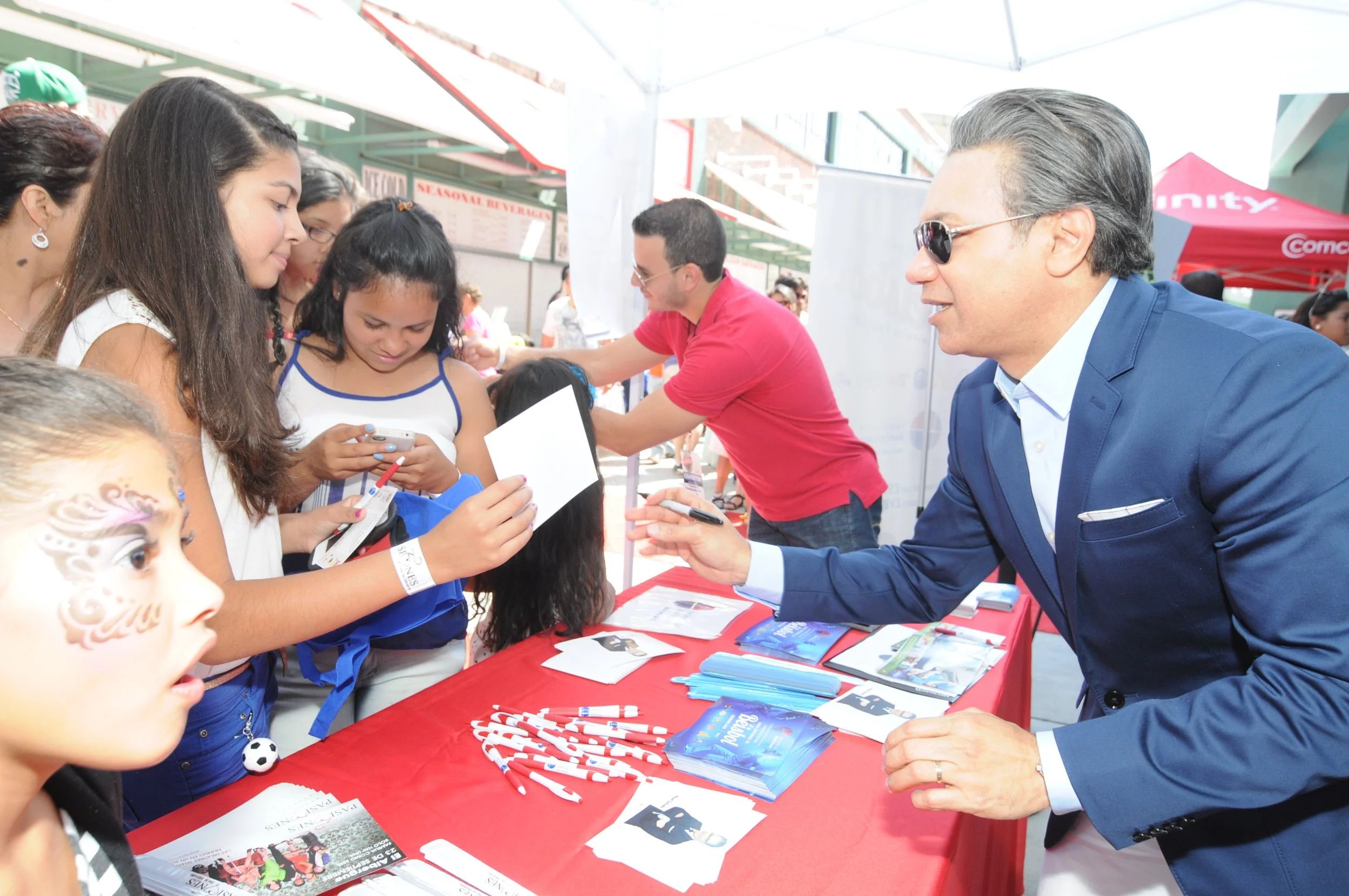 People at outdoor event booth exchanging flyers and brochures, man in suit signing autographs.
