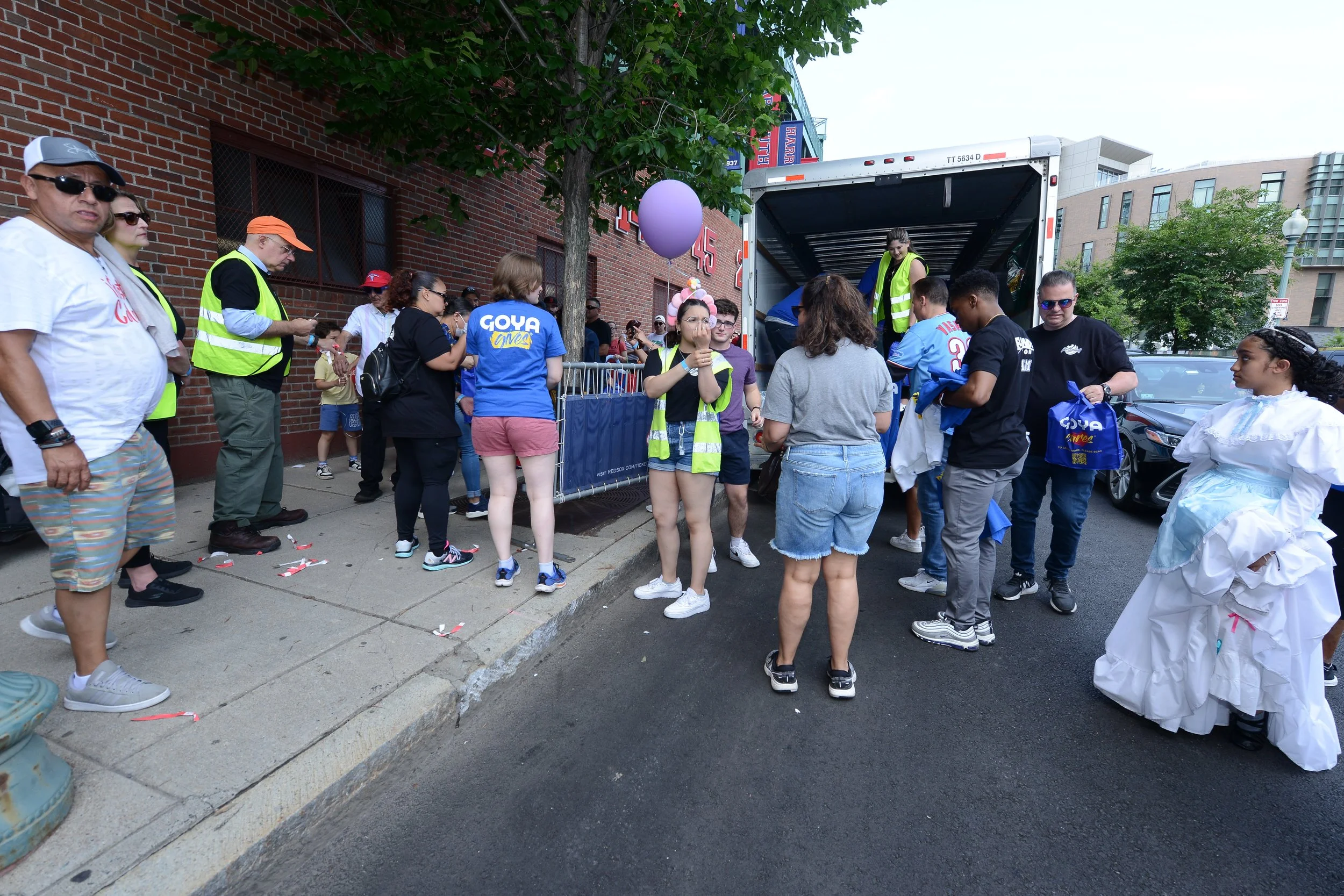 Group of people gathered near a truck on a street, some wearing 'Goya' shirts and vests, possibly distributing items. One person is holding a purple balloon. A child in a dress stands nearby. Brick building in the background.