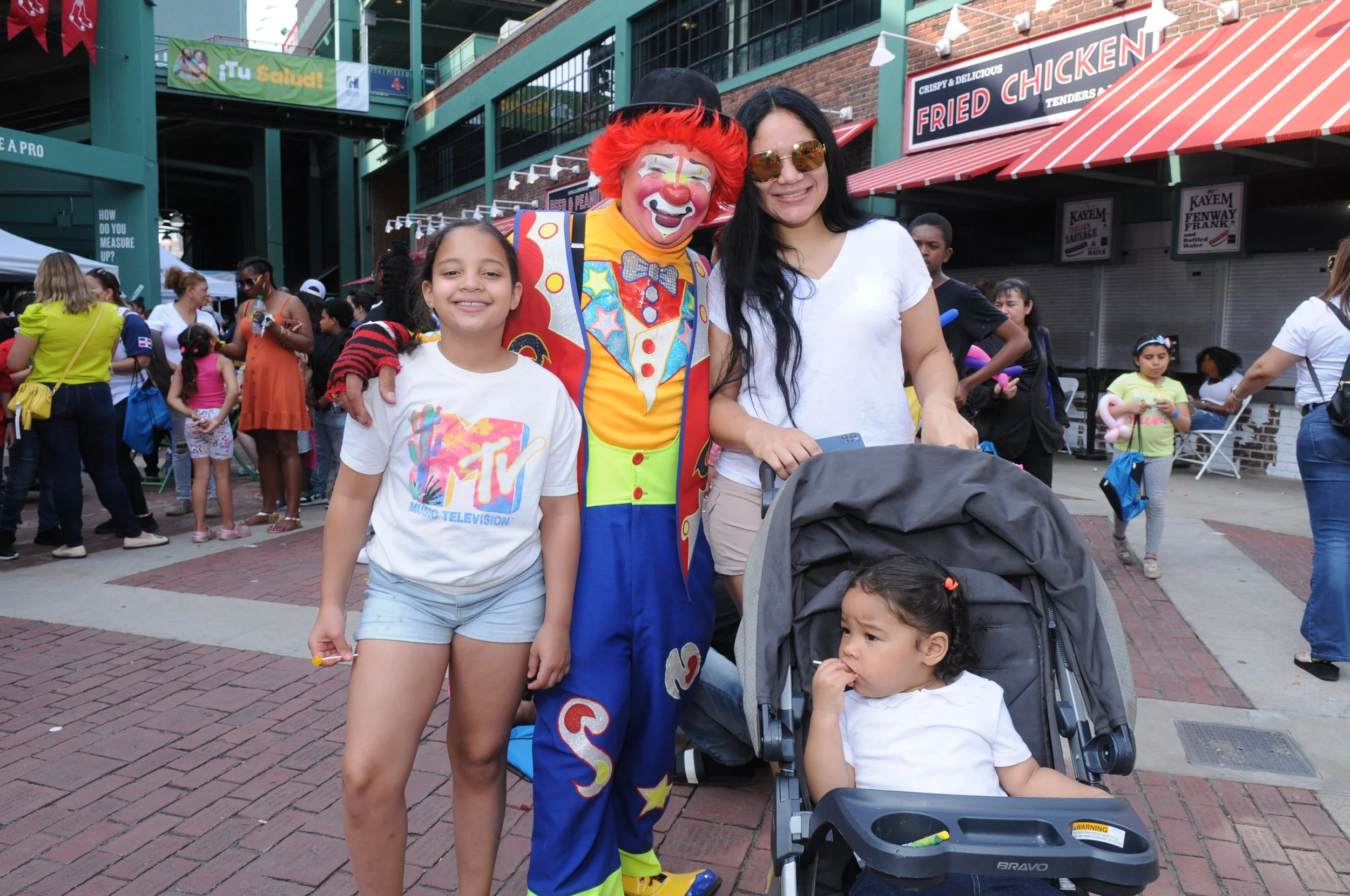 A clown posing with a woman and two children in front of a fried chicken stand at a crowded outdoor event.