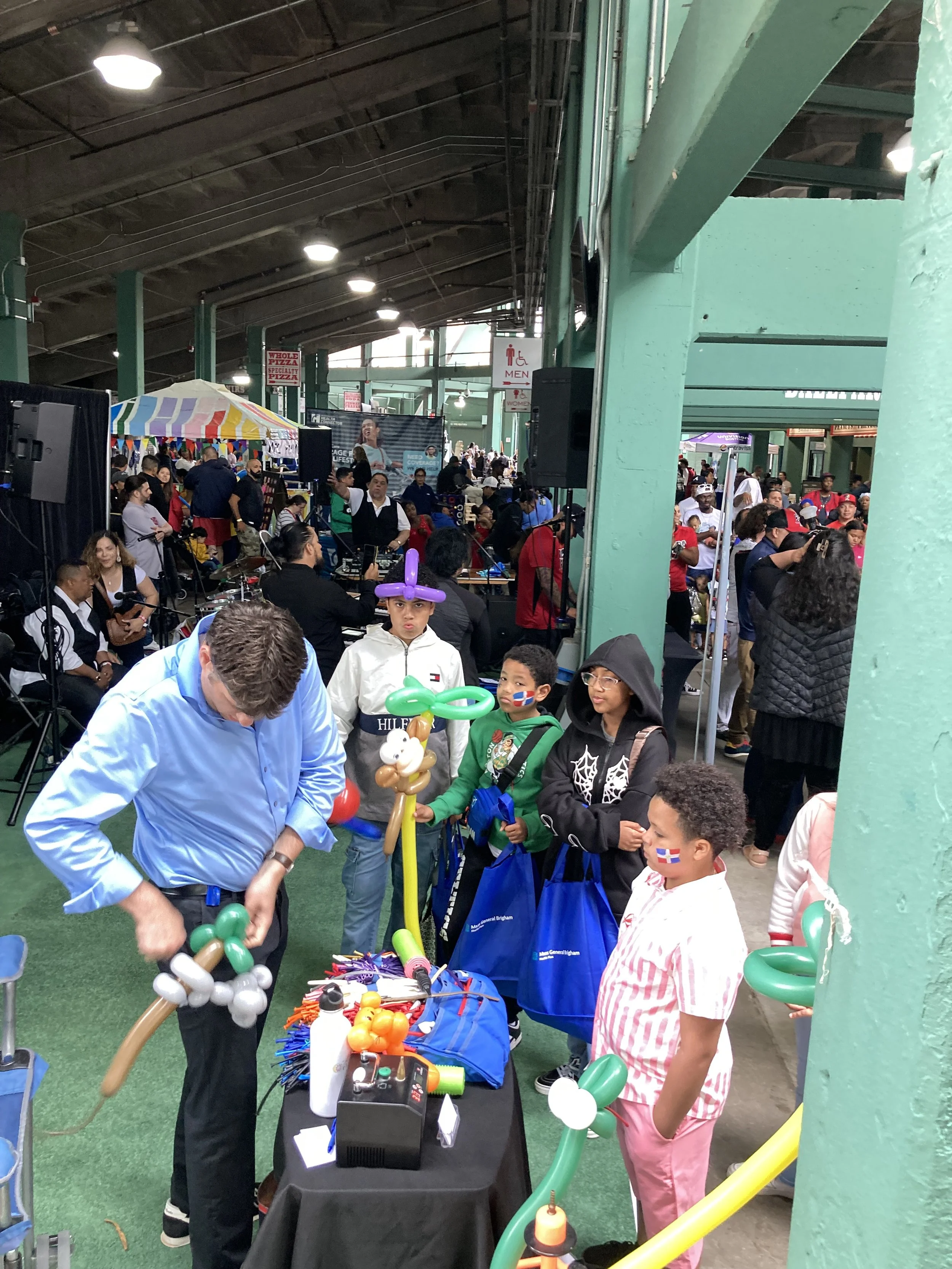 A balloon artist in a blue shirt making balloon animals at an indoor event with a crowd watching, including several children with face paint and colorful bags.