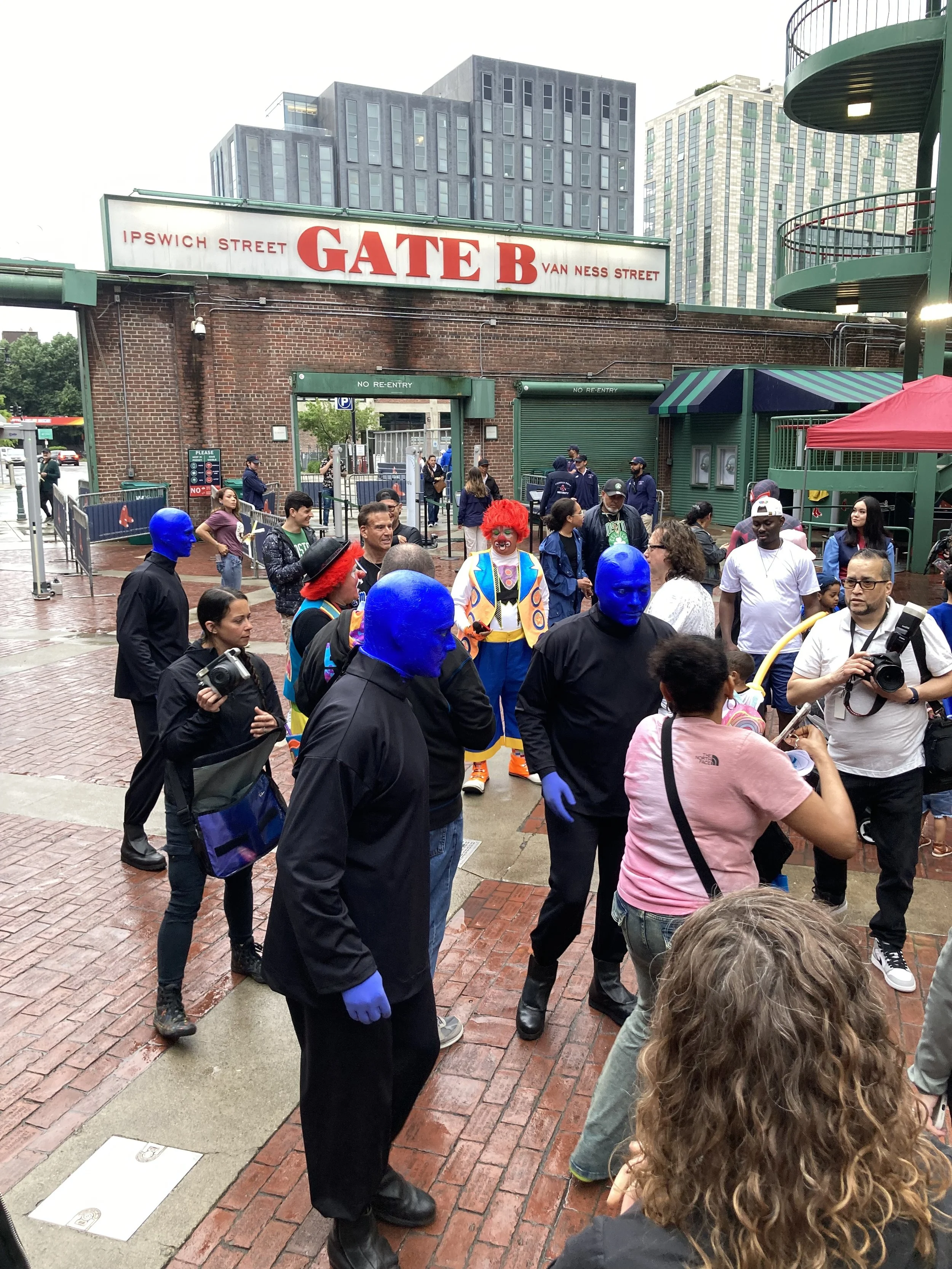 Crowd gathered outside a venue near "Gate B," featuring performers with painted blue faces and black outfits, and people dressed as clowns.