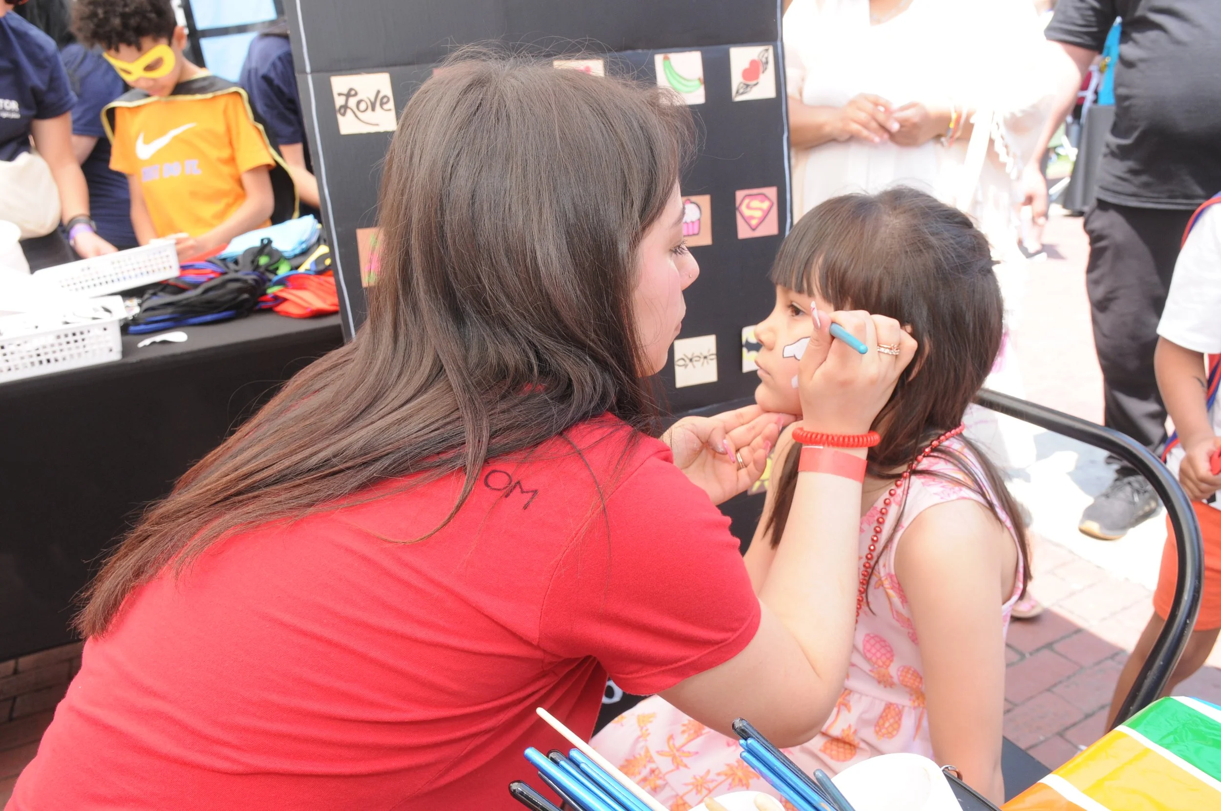 A woman painting a child's face at an outdoor event while a boy in a yellow mask stands nearby.