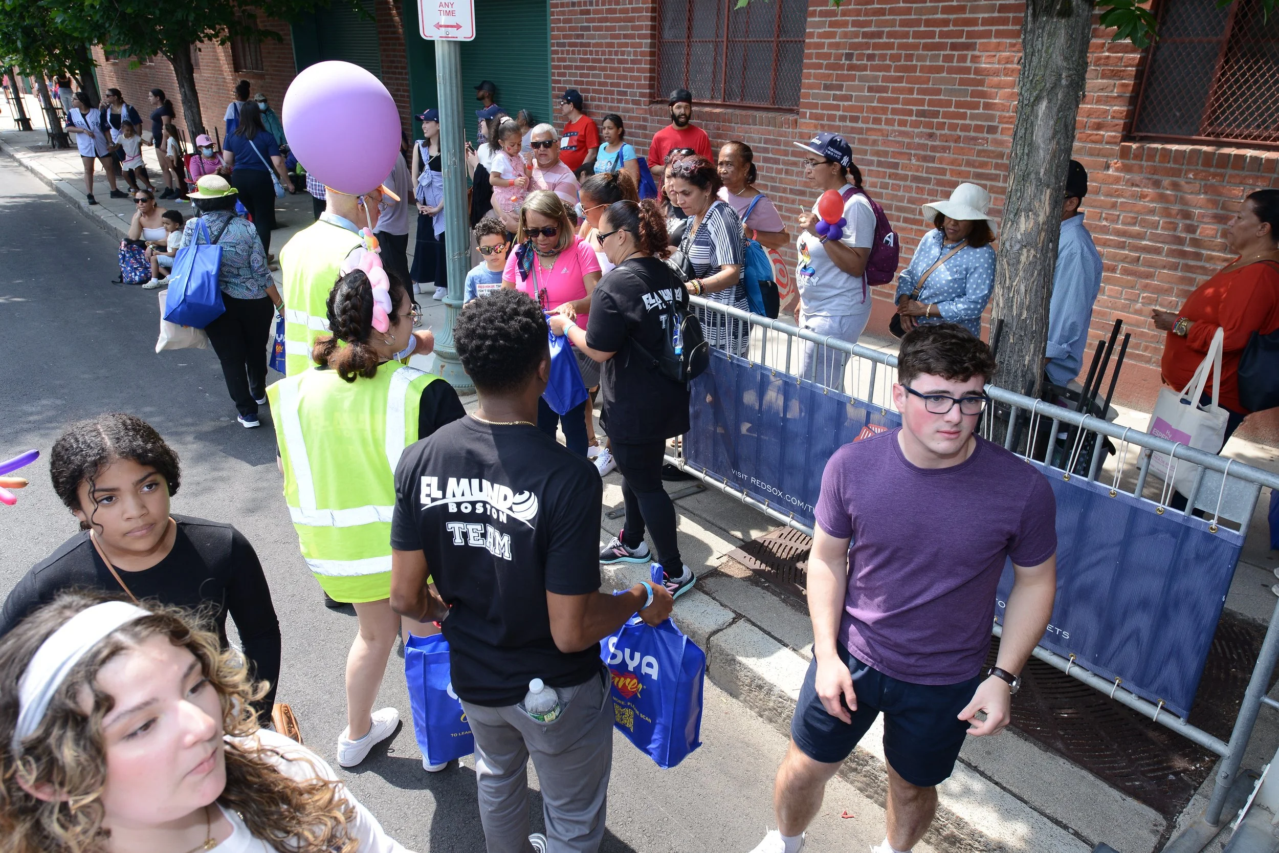 Crowd gathered on a street near a brick wall, some holding balloons and bags. People wearing "El Mundo Boston Team" shirts are distributing bags. Other individuals are lined up along a temporary fence.