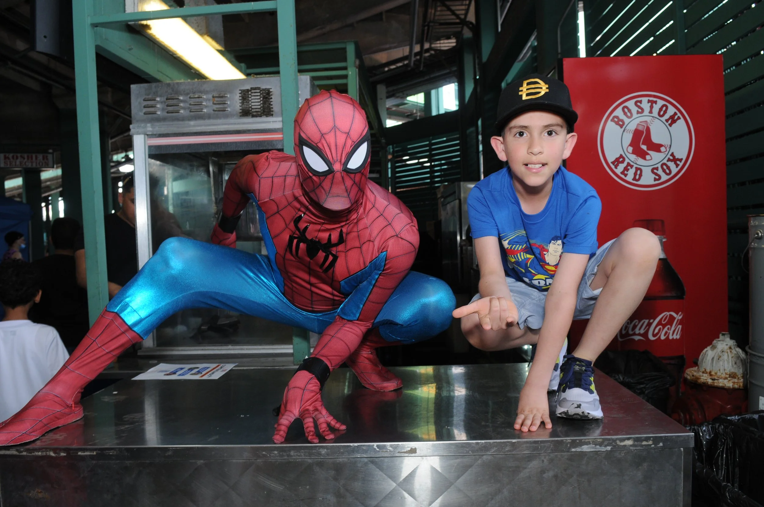 Person in Spider-Man costume posing with a child wearing a blue shirt and baseball cap, in front of a Boston Red Sox logo.