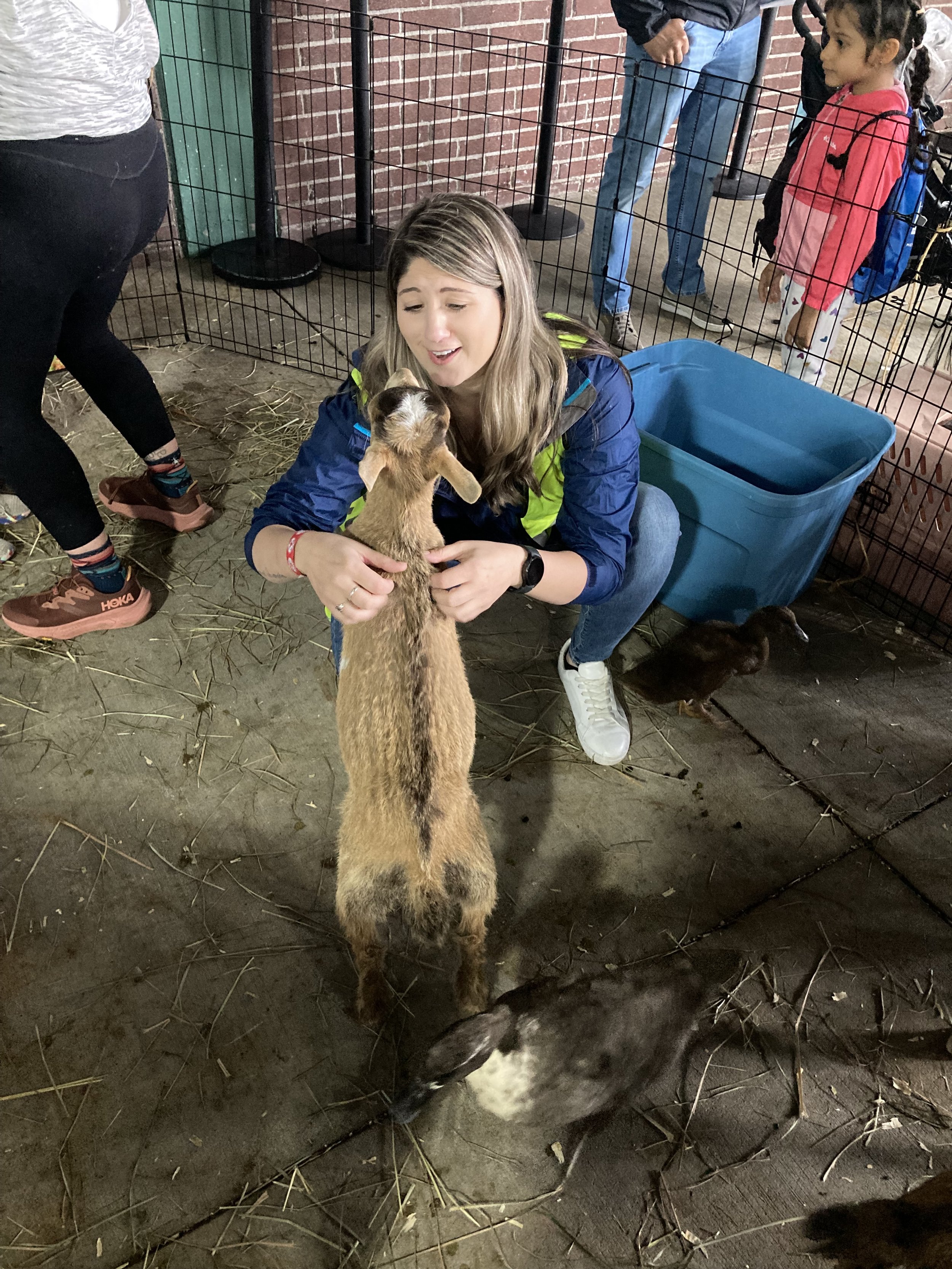 A woman crouching down petting a small goat in an enclosure, with a child standing nearby watching."