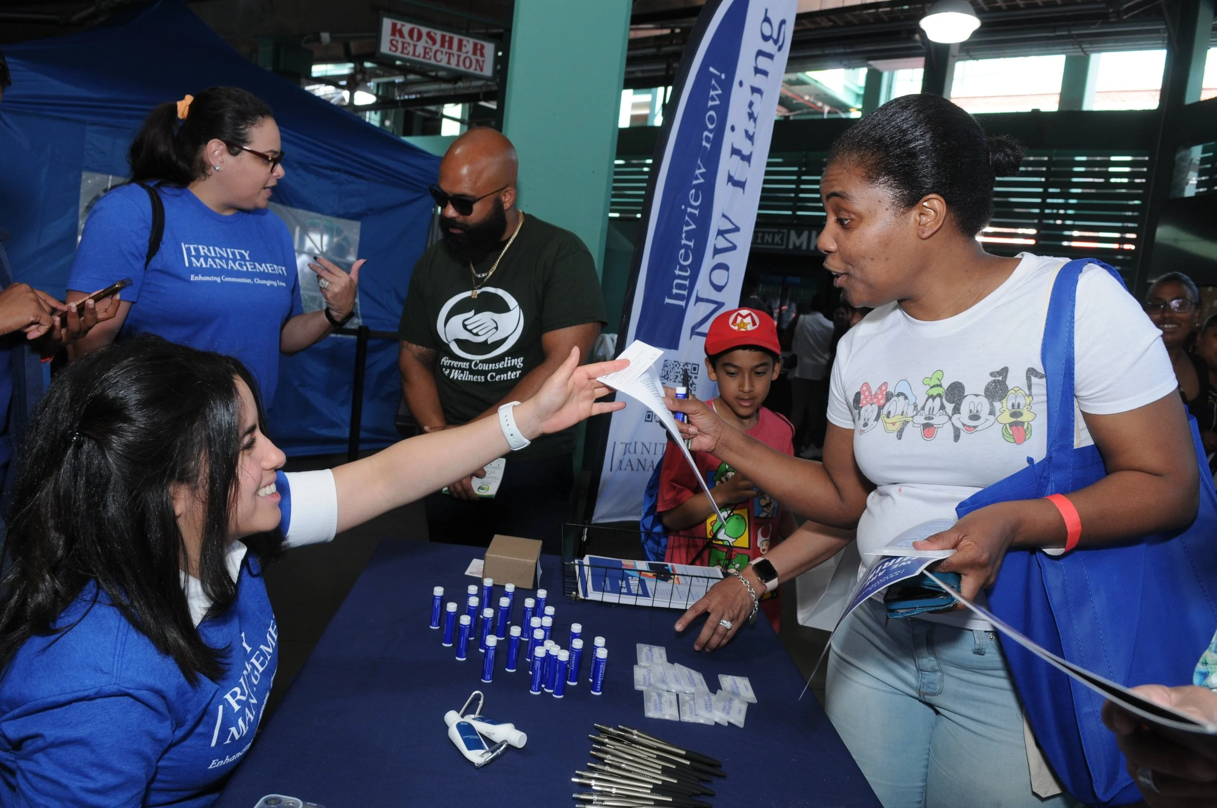 People interacting at a booth with promotional items, including pens and tubes, a "Now Hiring" banner, and a person handing a flyer to another person.
