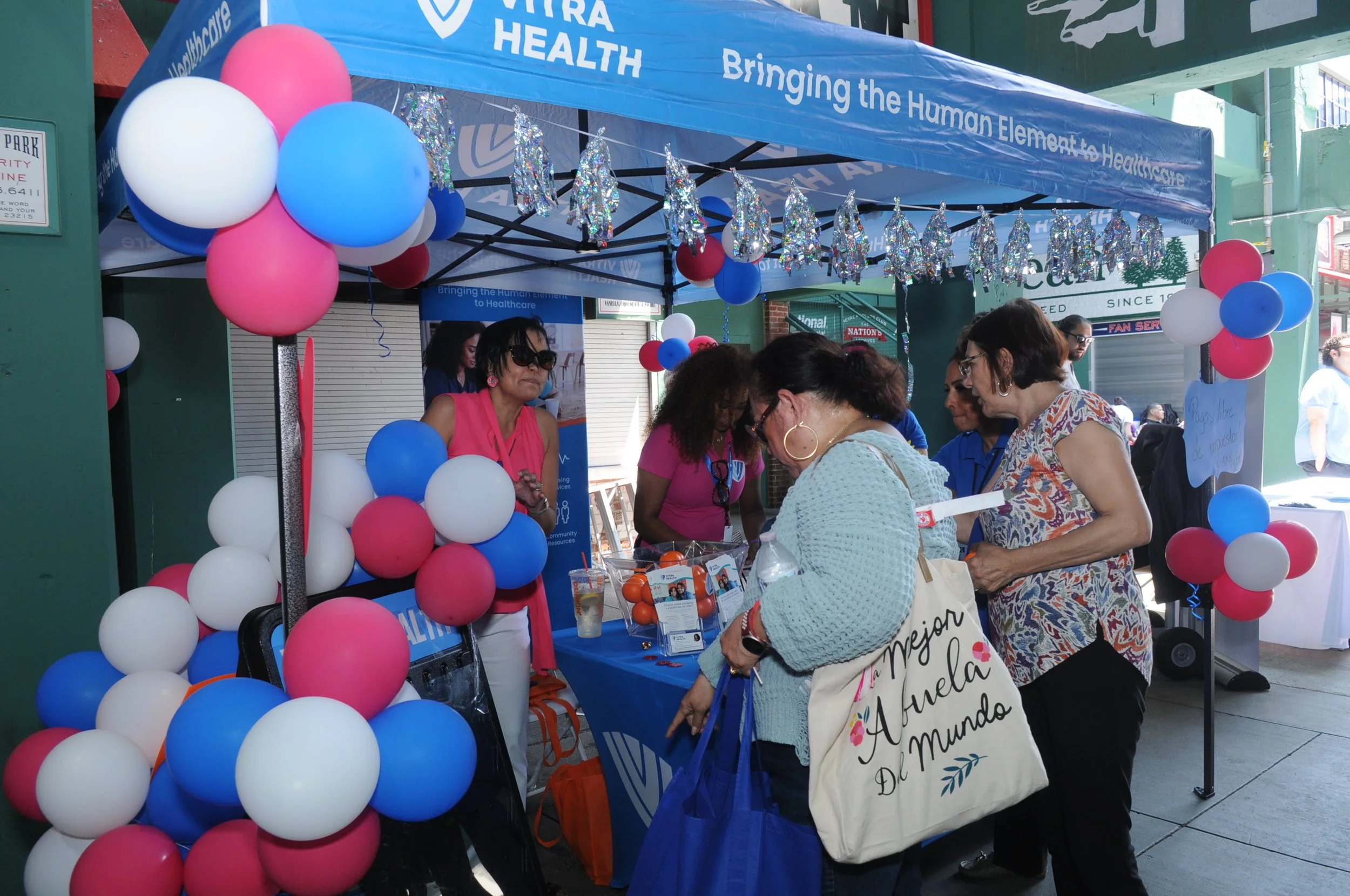 People interacting at a health booth under a blue tent, decorated with red, white, and blue balloons.