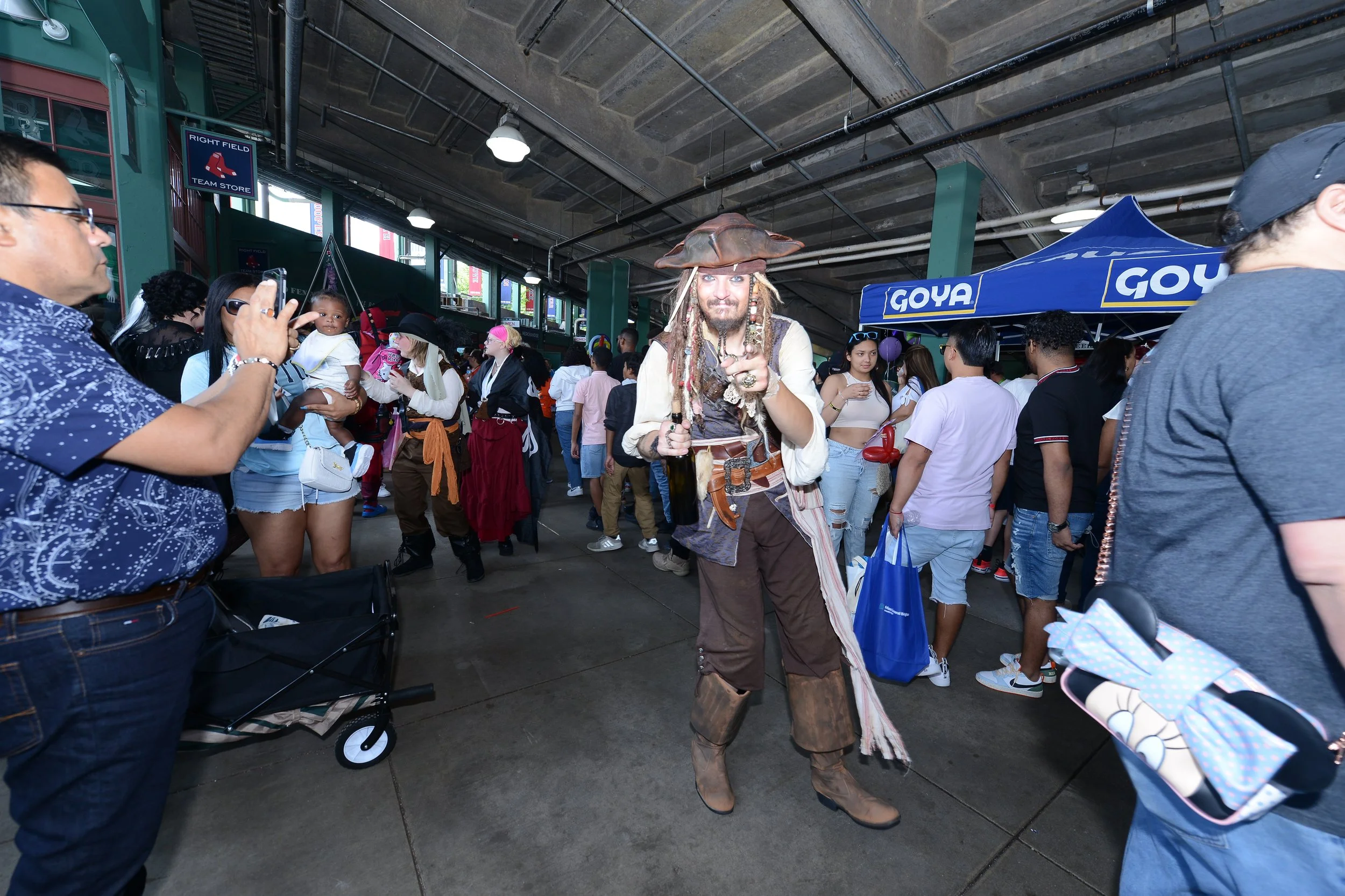 People dressed in pirate costumes at an indoor event, with a man in a pirate outfit posing for the camera. A crowd surrounds a Goya-branded tent.