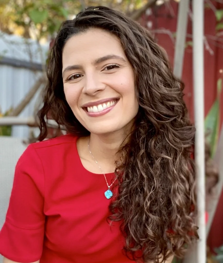 Smiling woman with curly brown hair wearing a red top and a silver necklace with a blue pendant, outdoors with a red fence and greenery in the background.
