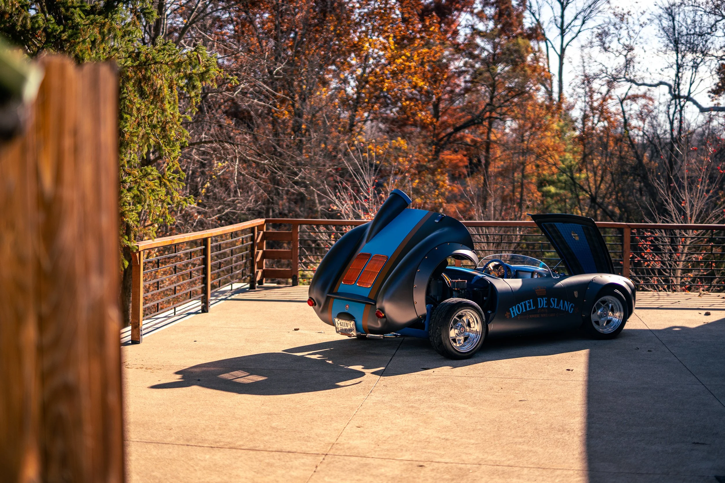 A small, vintage-style car painted in dark blue with a tan stripe in the middle, parked on a concrete surface with a shadow cast to the left. The car has an open, curved body and a sign on the side that reads 'HOTEL DE SLANG'. It's outdoors on a deck or balcony with a wooden railing, surrounded by trees with autumn foliage in the background.