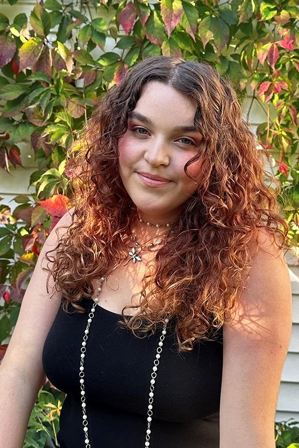 A young woman with curly brown hair and fair skin standing outdoors in front of a colorful plant with red, green, and pink leaves, wearing a black sleeveless top and layered necklaces with a flower pendant.