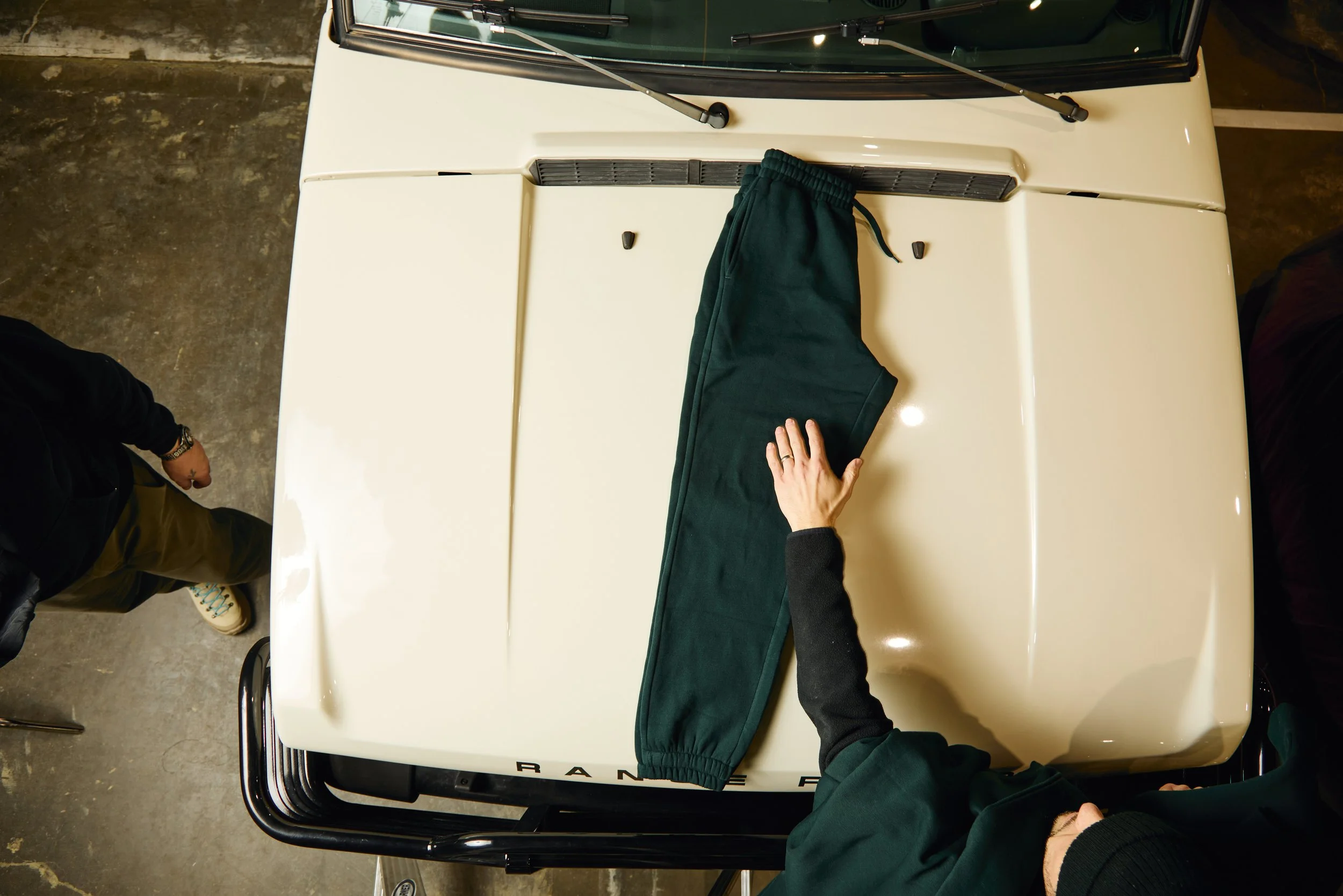 Overhead view of a person folding dark green pants on top of the hood of a white Range Rover vehicle in a parking garage. The person's hands are visible, and they are wearing a dark long-sleeve shirt. Another person, wearing a black jacket and beige shoes, is standing nearby.