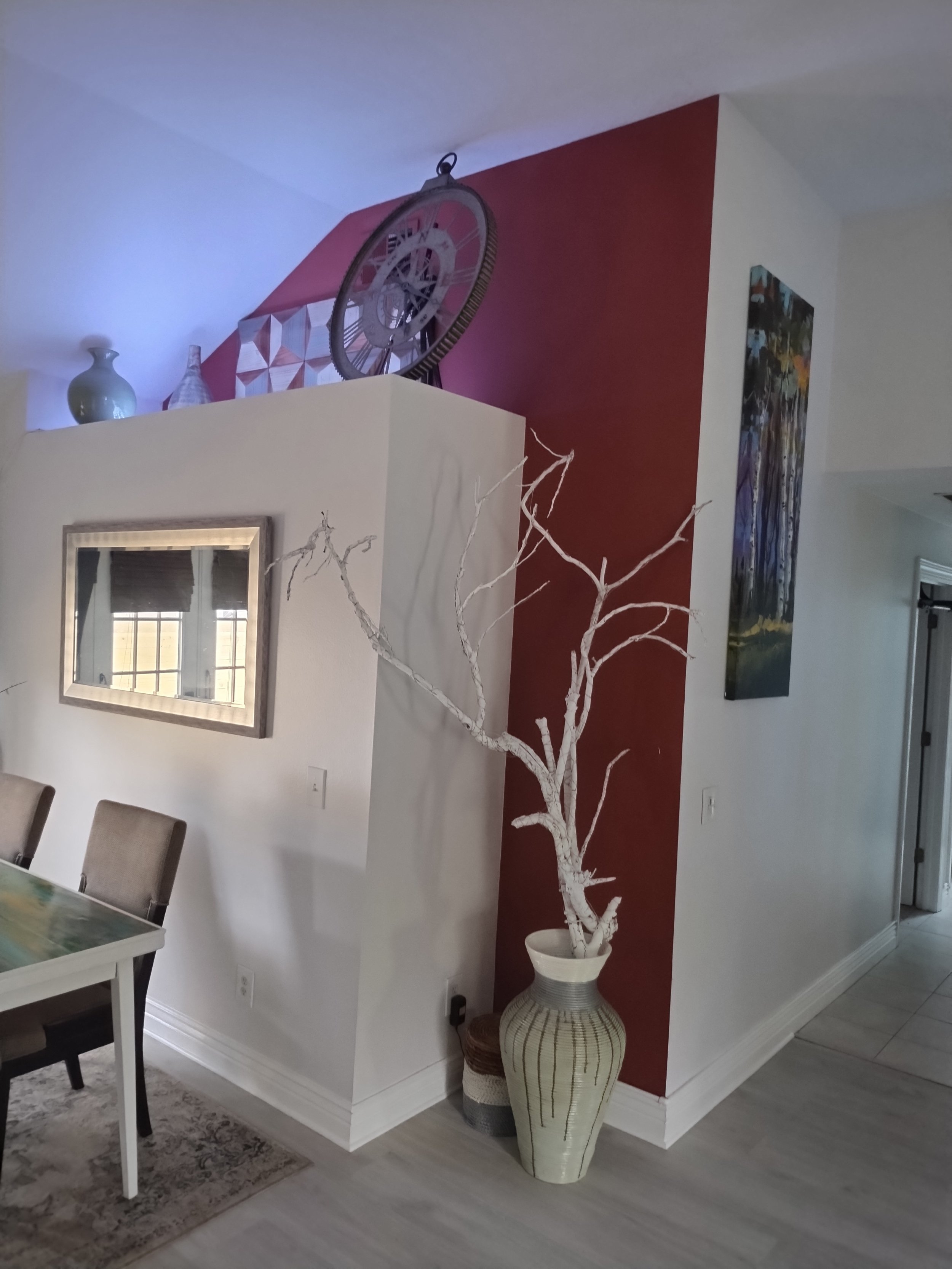 Interior of a modern dining room with a tall white vase containing a decorative branch, a wall with red and white sections, various decor items on a shelf, and beige chairs around a table.