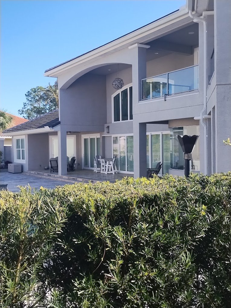 Modern gray house with large windows and balconies, featuring a patio with outdoor furniture, surrounded by green shrubs.