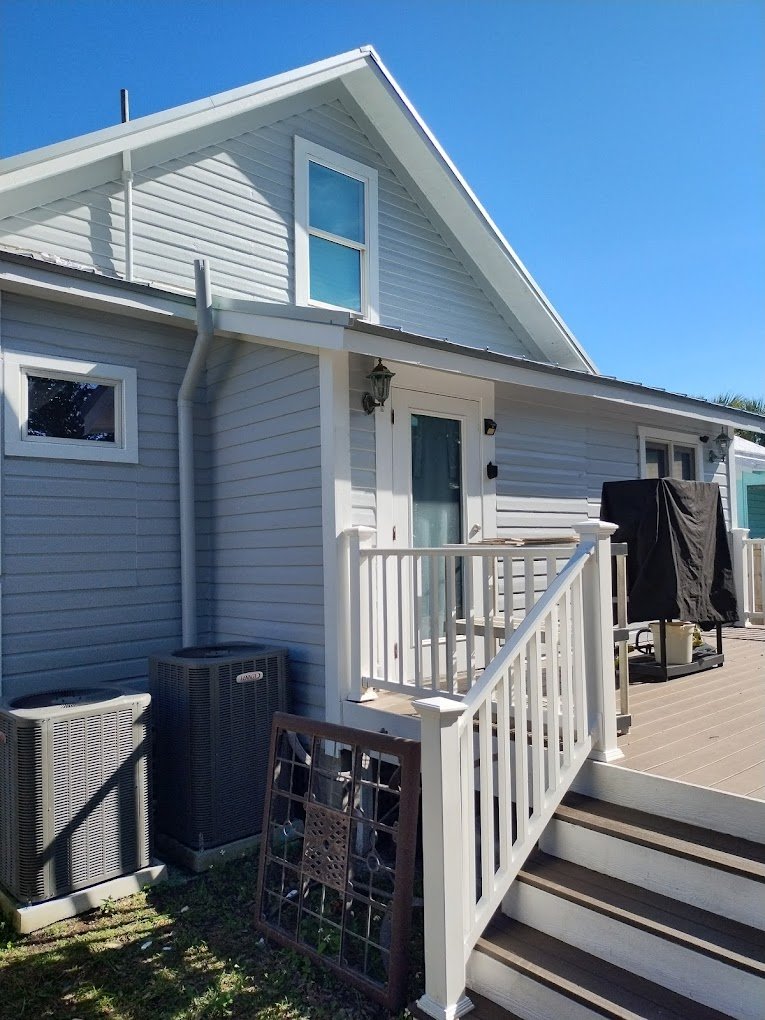 Exterior of a light gray house with white trim, featuring a small porch with white railings and stairs. Air conditioning units are visible on the left, and a covered grill on a wooden deck on the right. A decorative wrought iron piece leans against t