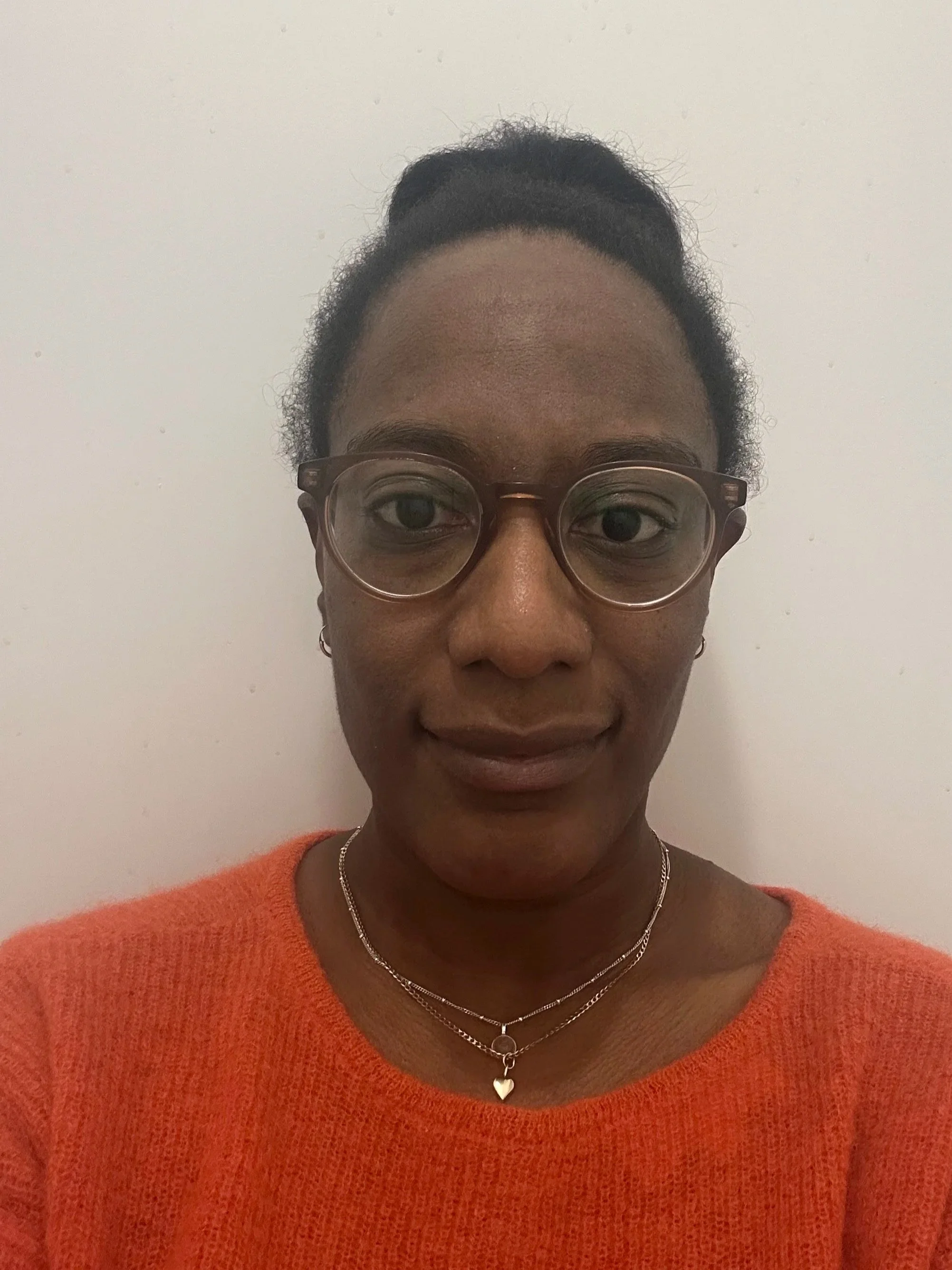 A woman with dark skin and glasses takes a selfie in front of a plain white wall. She is wearing an orange sweater and layered necklaces, including a heart-shaped pendant.