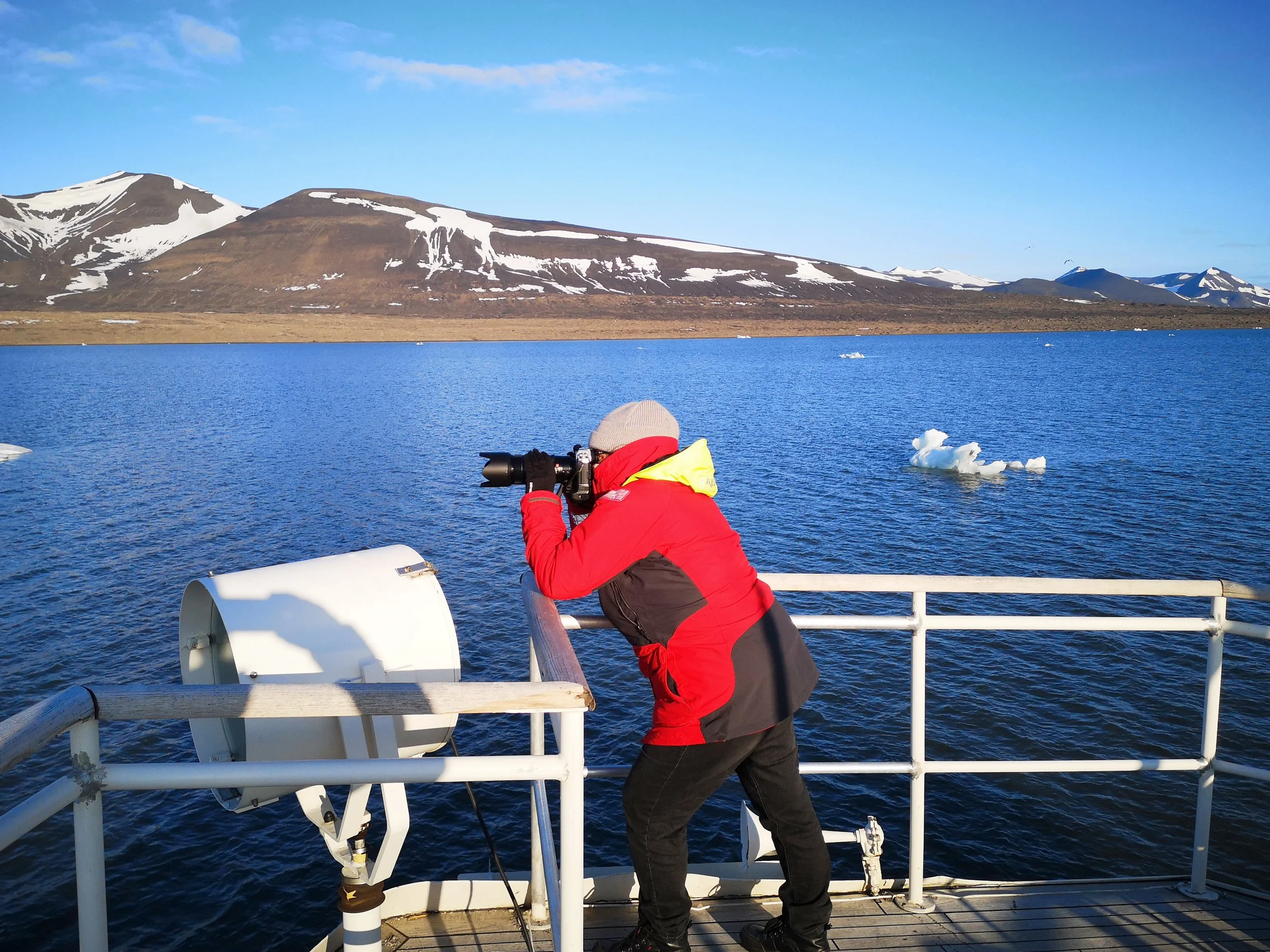 A researcher in red and black winter clothing, wearing a beige beanie, is looking through a camera with snow-capped mountains in the background.