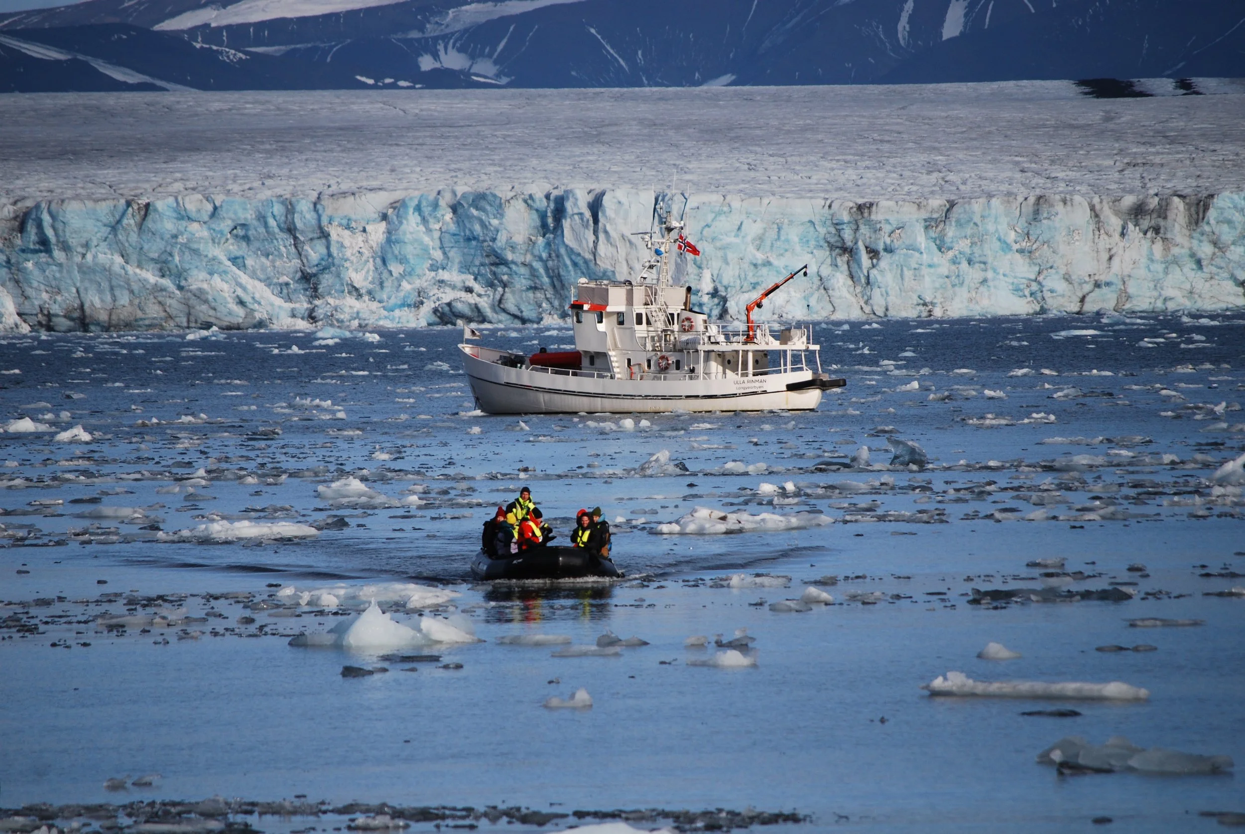 A group of people in a small inflatable boat navigating icy waters near a large glacier, with a research vessel anchored nearby in a polar environment.