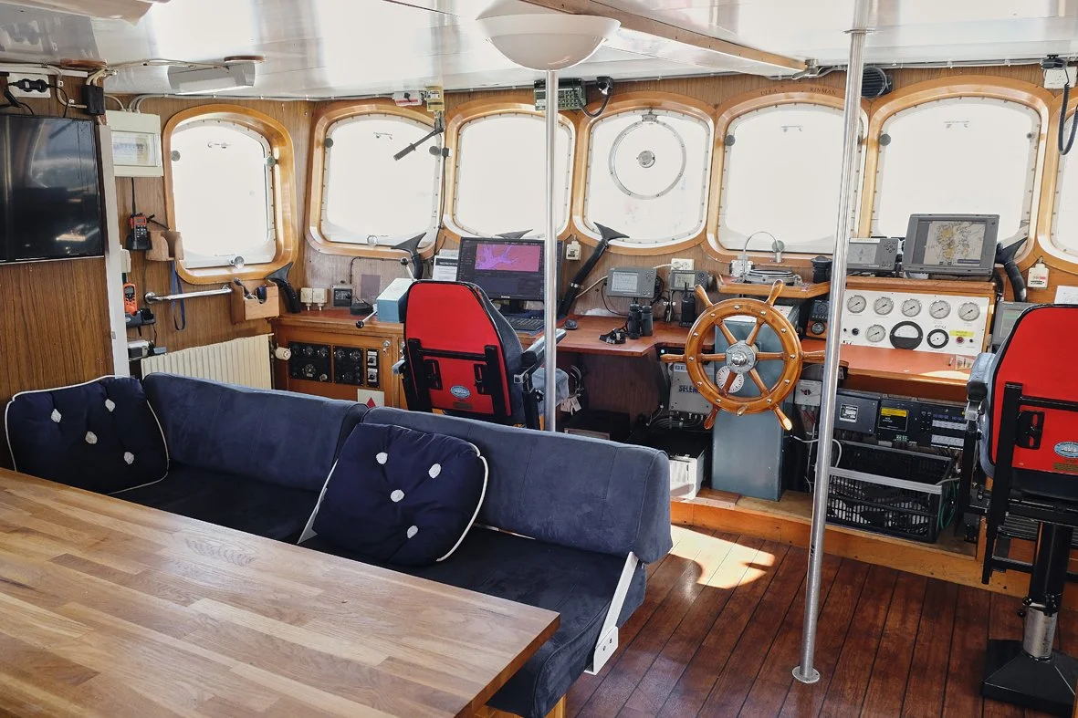 Interior of a ship's bridge with navigational equipment, steering wheel, computer screens, and a sofa with cushions.