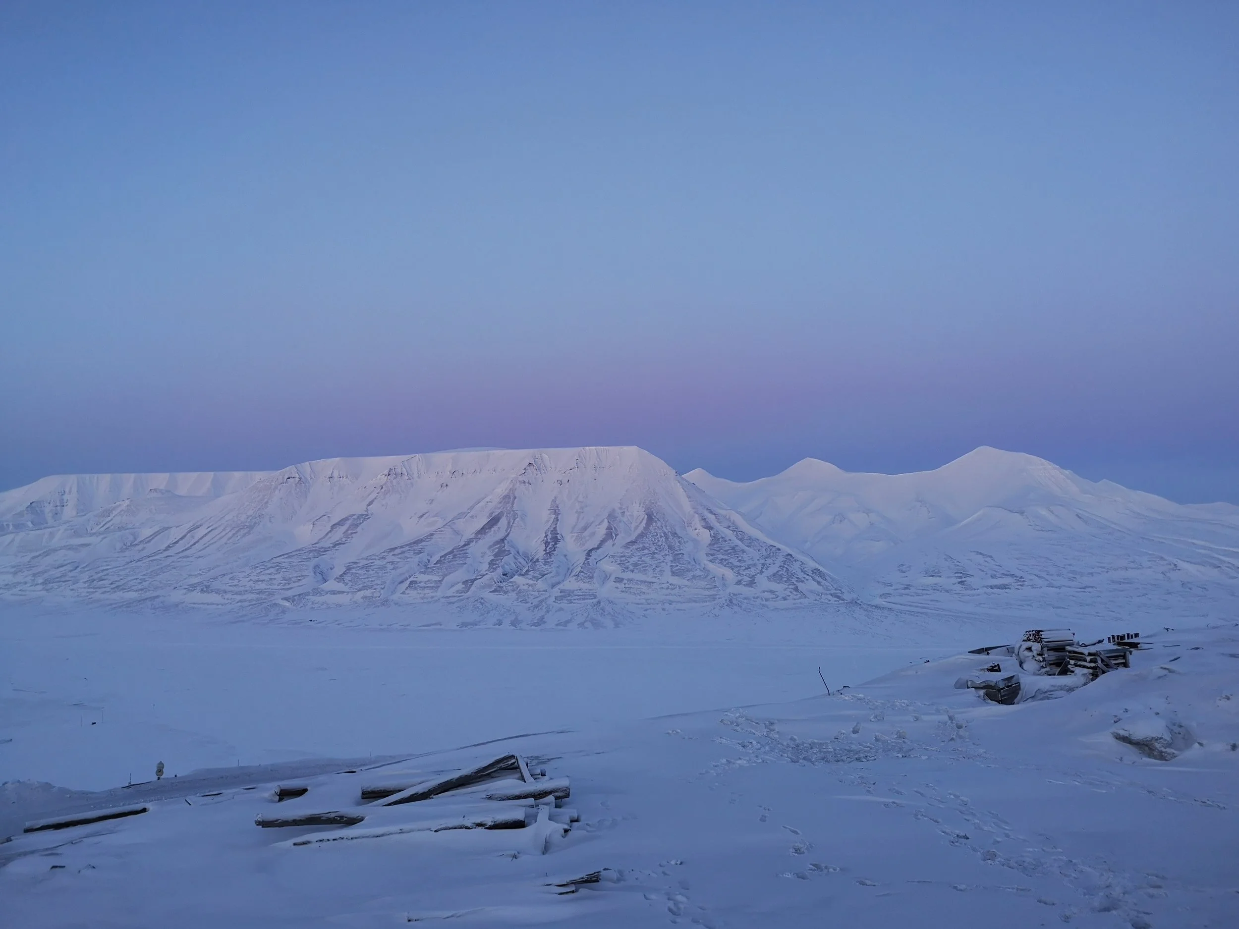 Snow-covered mountain range under a clear blue sky, with a few debris and structures in the foreground.