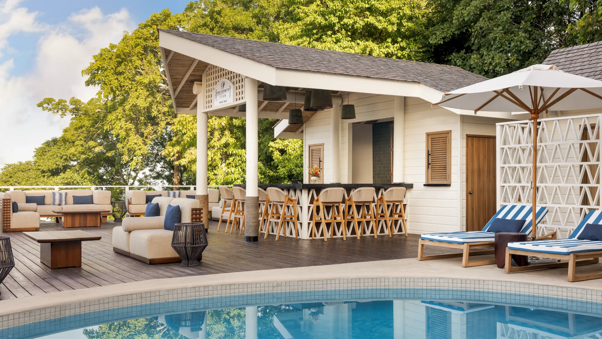 Outdoor pool area with lounge chairs, umbrellas, and a bar with barstools, surrounded by lush green trees.